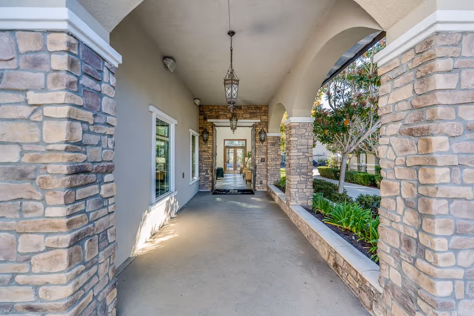 Covered stone walkway with arches and lanterns along a senior living facility entrance.