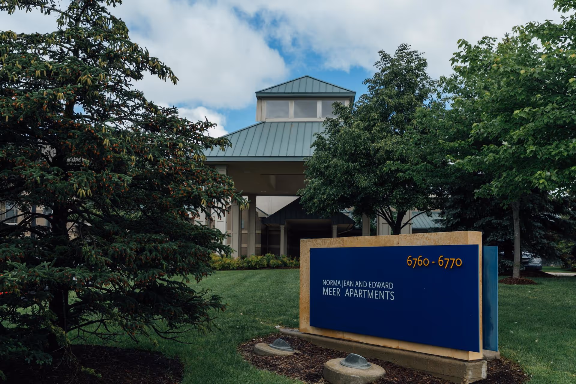 Front entrance of an apartment building with a blue sign reading 'Norma Jean and Edward Meer Apartments' and the address 6760-6770, surrounded by trees and lawn.