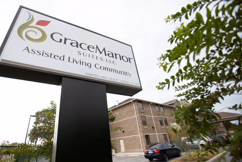 A large 'Grace Manor Suites' sign stands in front of a brick assisted living building with parked cars and trees.