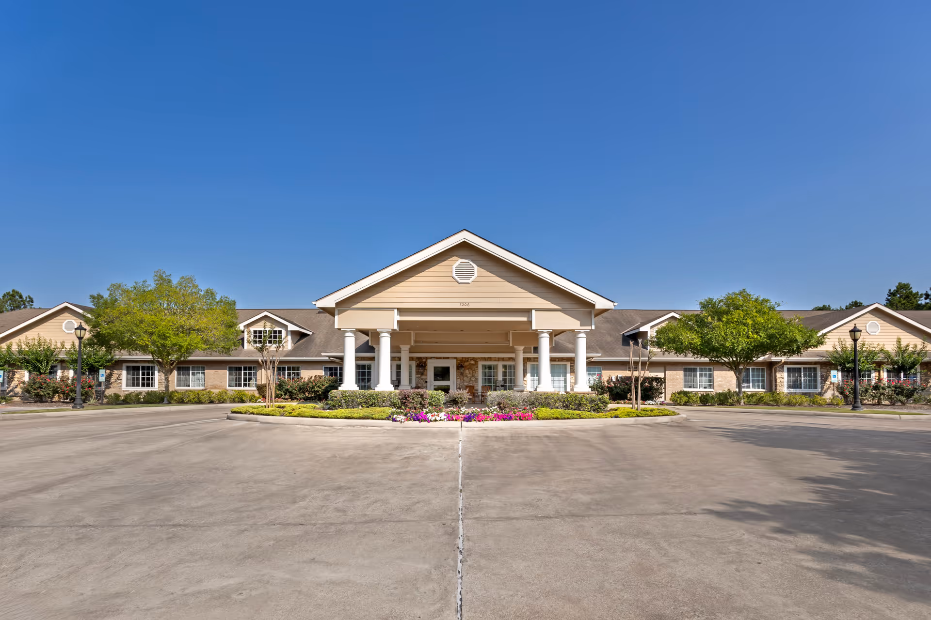 Front exterior view of Cinco Ranch Alzheimer's Special Care Center, a single-story building with a covered entrance supported by white columns, surrounded by landscaped greenery and flowers under a clear blue sky.
