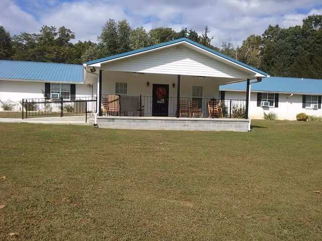 Single-story white building with blue roof and a covered porch with rocking chairs.