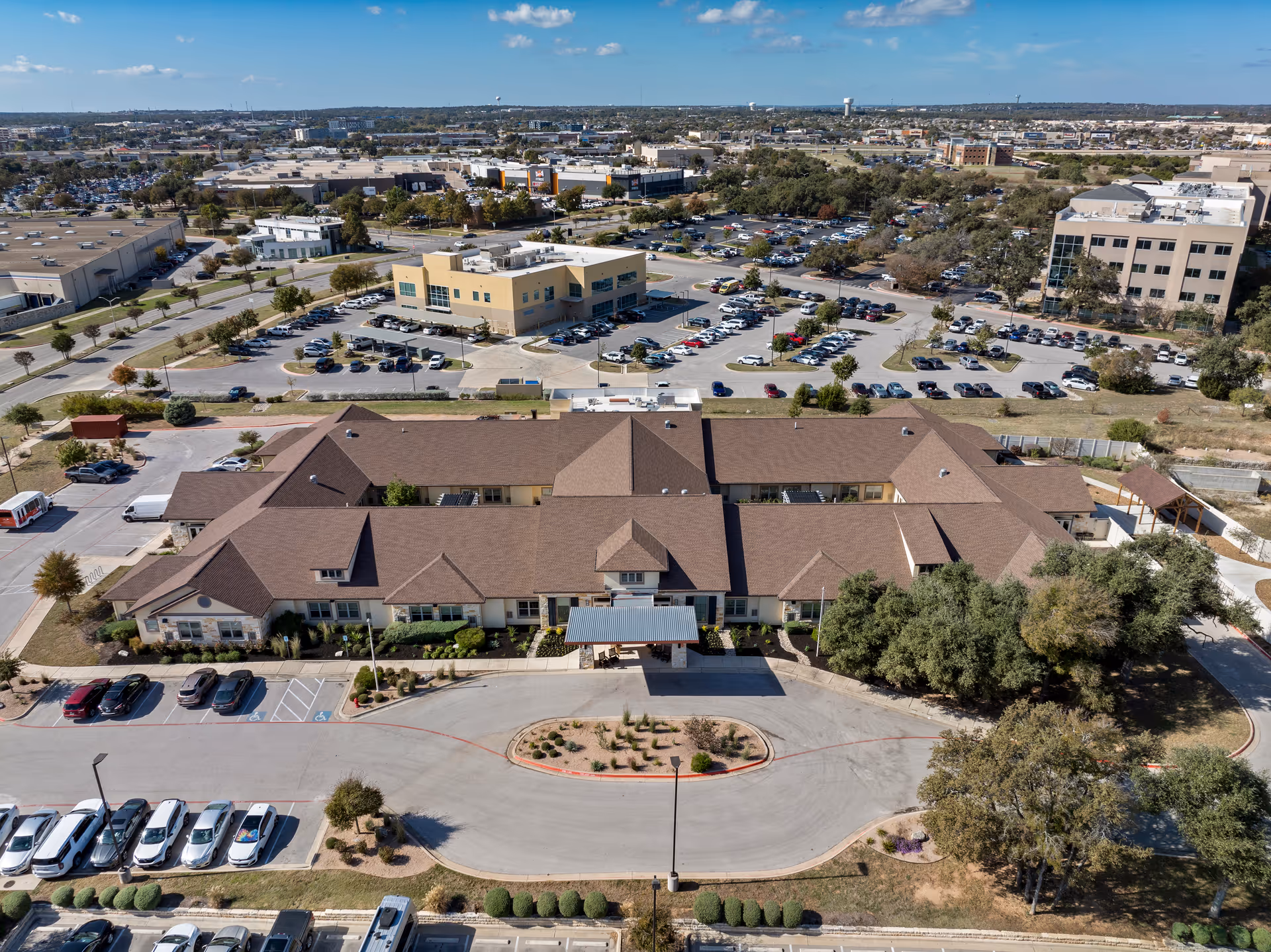 Aerial view of The Monarch at Cedar Park senior living facility showing a large building with a brown roof surrounded by parking lots, trees, and other commercial buildings in the background under a partly cloudy sky.