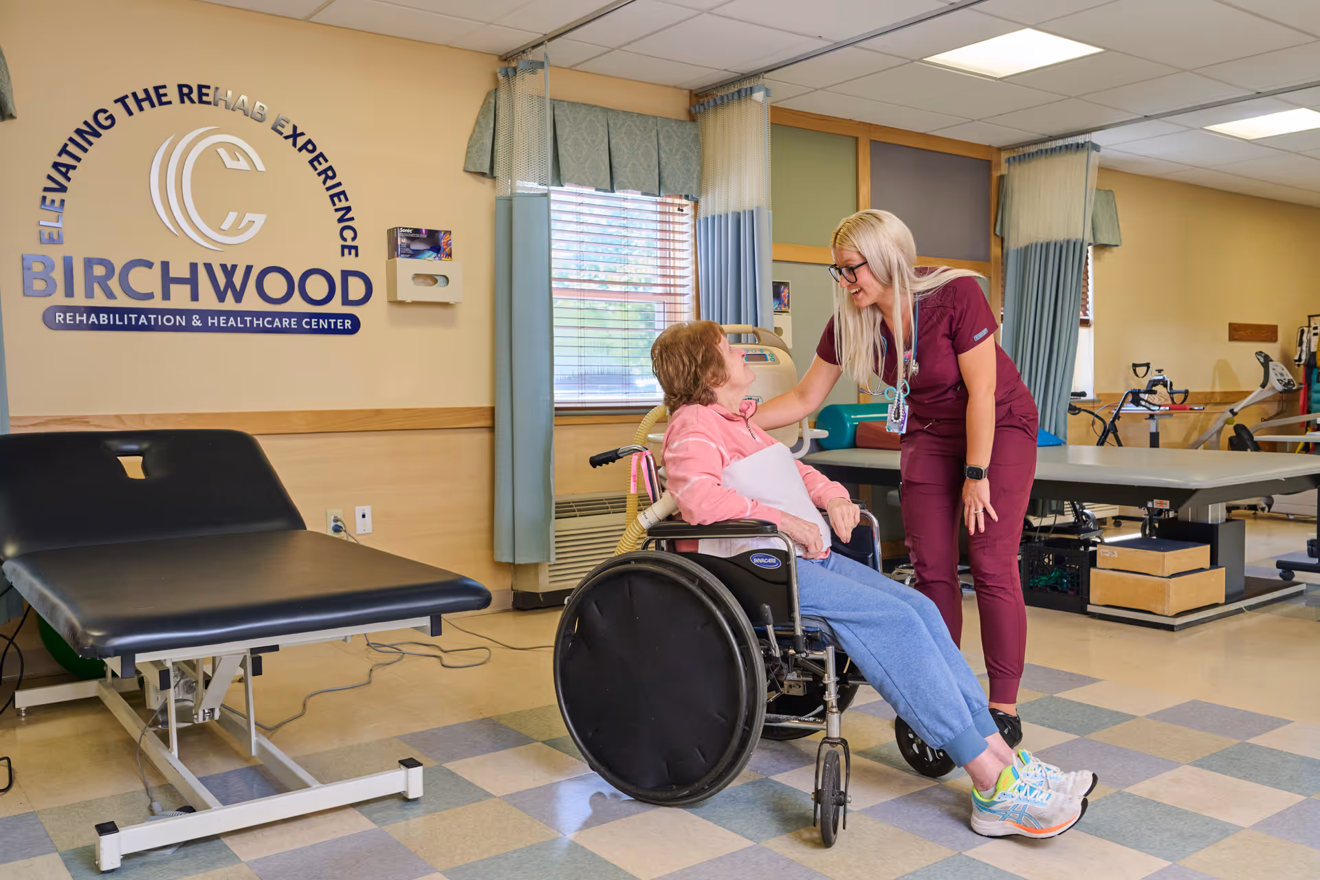 A healthcare worker in maroon scrubs is smiling and interacting with an elderly woman seated in a wheelchair inside a rehabilitation room. The room has medical equipment, a treatment table, and windows with curtains. The wall displays the Birchwood Rehabilitation & Healthcare Center logo and slogan.