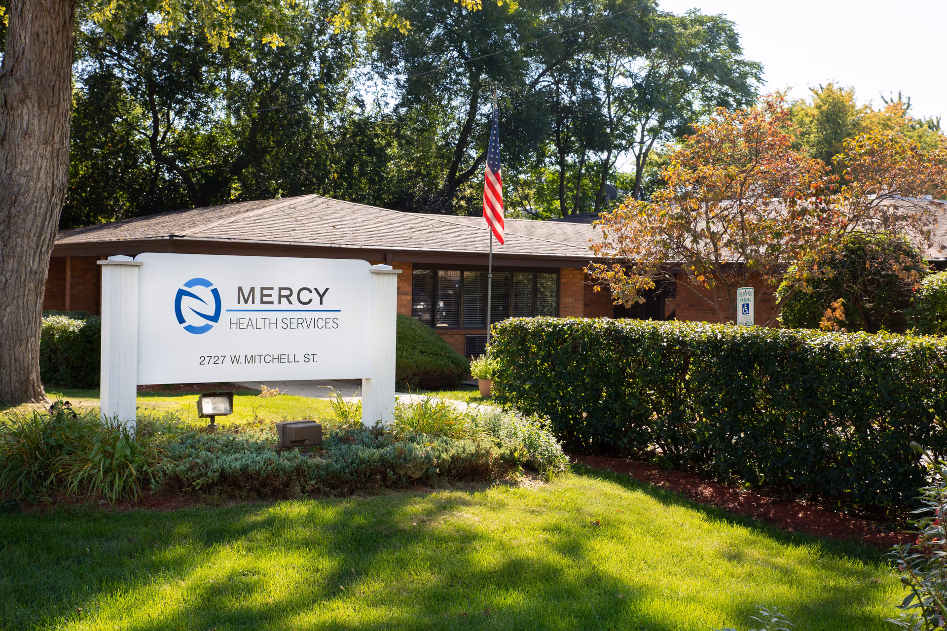 Mercy Health Services sign in front of a low brick building with trees and a flag.