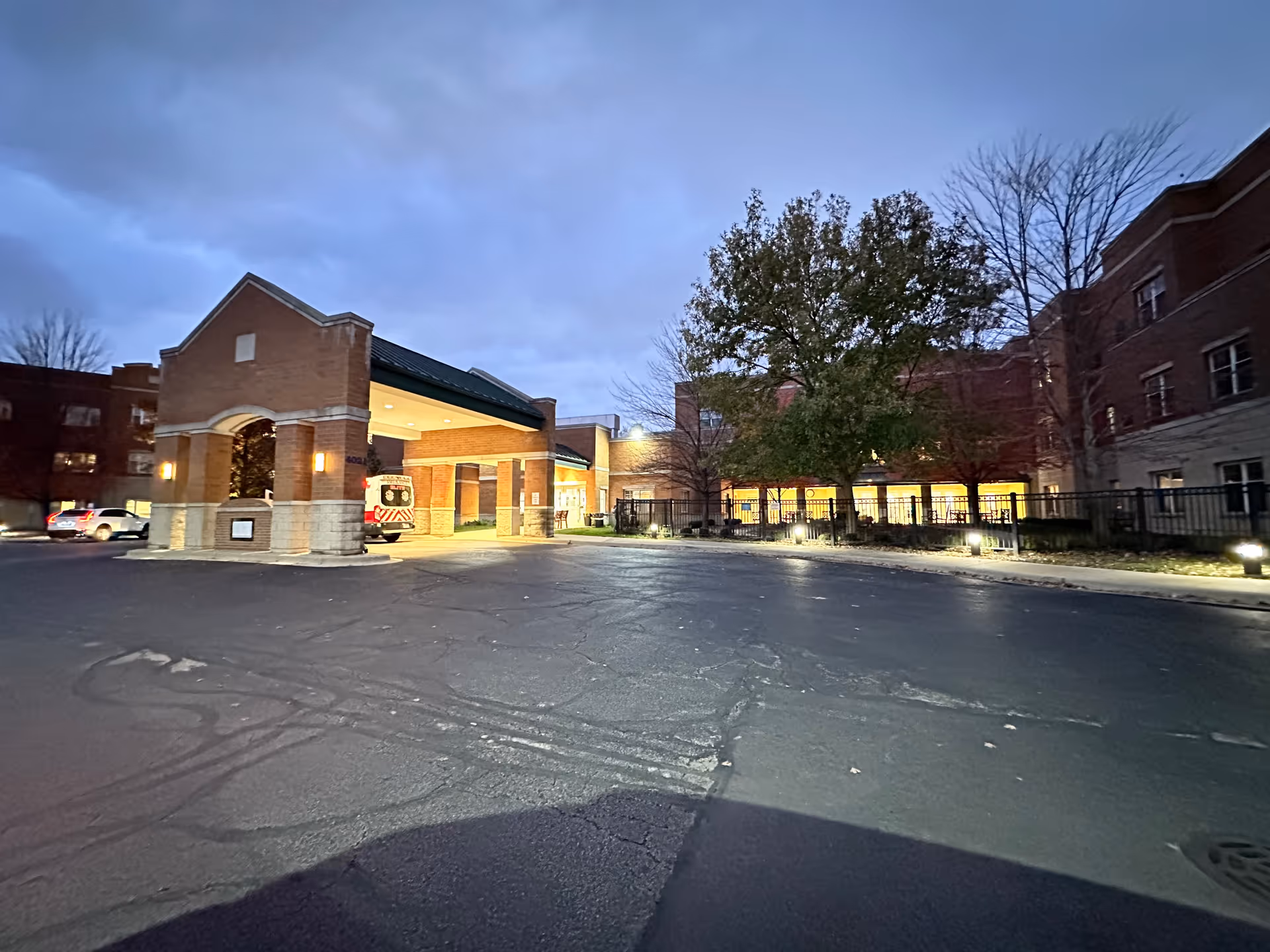 Dusk view of the brick entrance and porte-cochere of a senior living facility with lit windows, trees, and vehicles in the driveway.