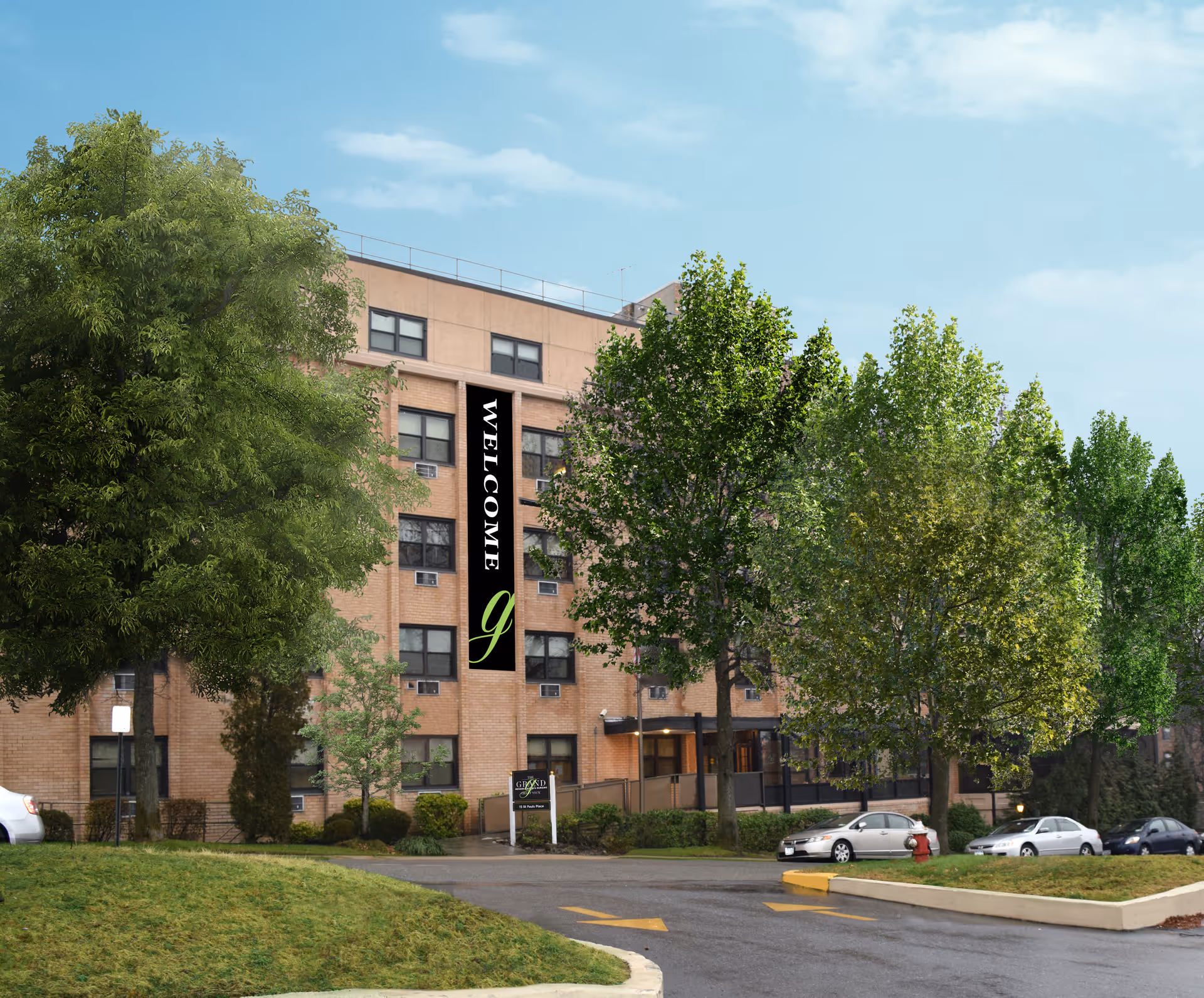 Exterior view of a multi-story brick building with several windows, surrounded by green trees and a parking area with cars. A vertical banner on the building reads 'WELCOME' with a stylized letter 'g' below it. The sky is clear with some clouds.