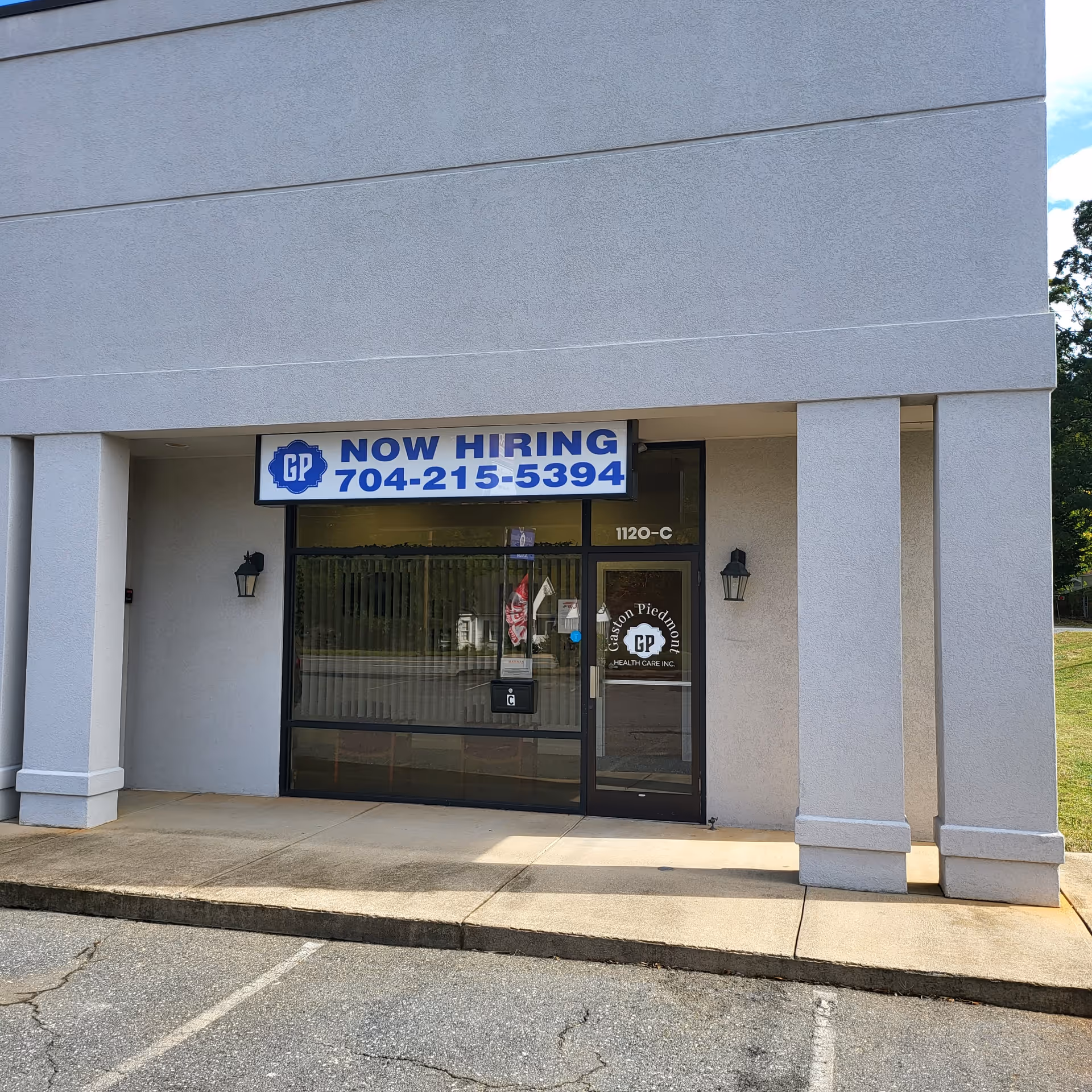 Front entrance of a grey building with a 'Now Hiring' sign and glass doors