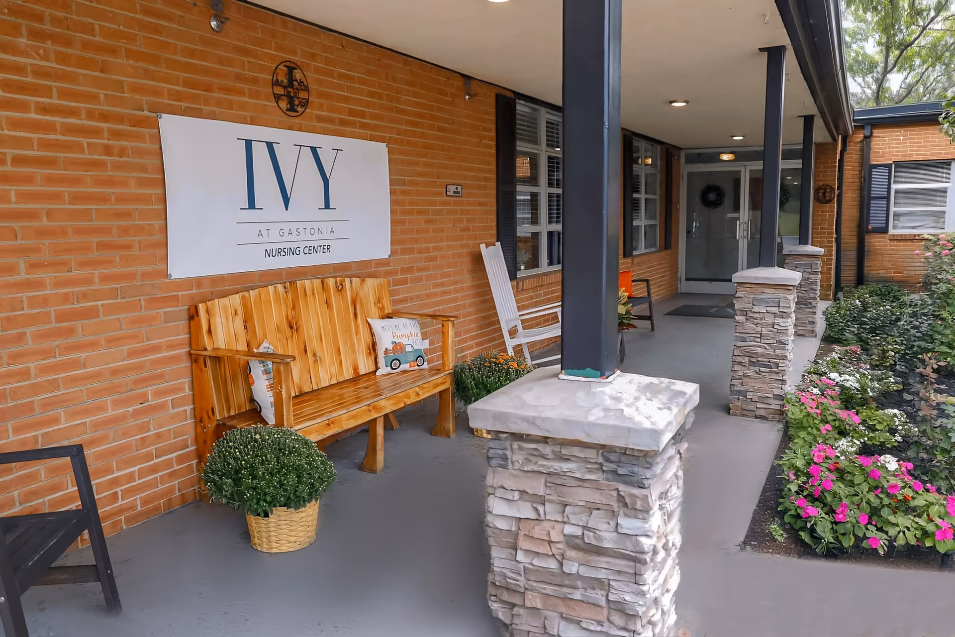 Covered brick entrance porch with a wooden bench, rocking chairs, potted plants, and a sign for The Ivy at Gastonia nursing center.