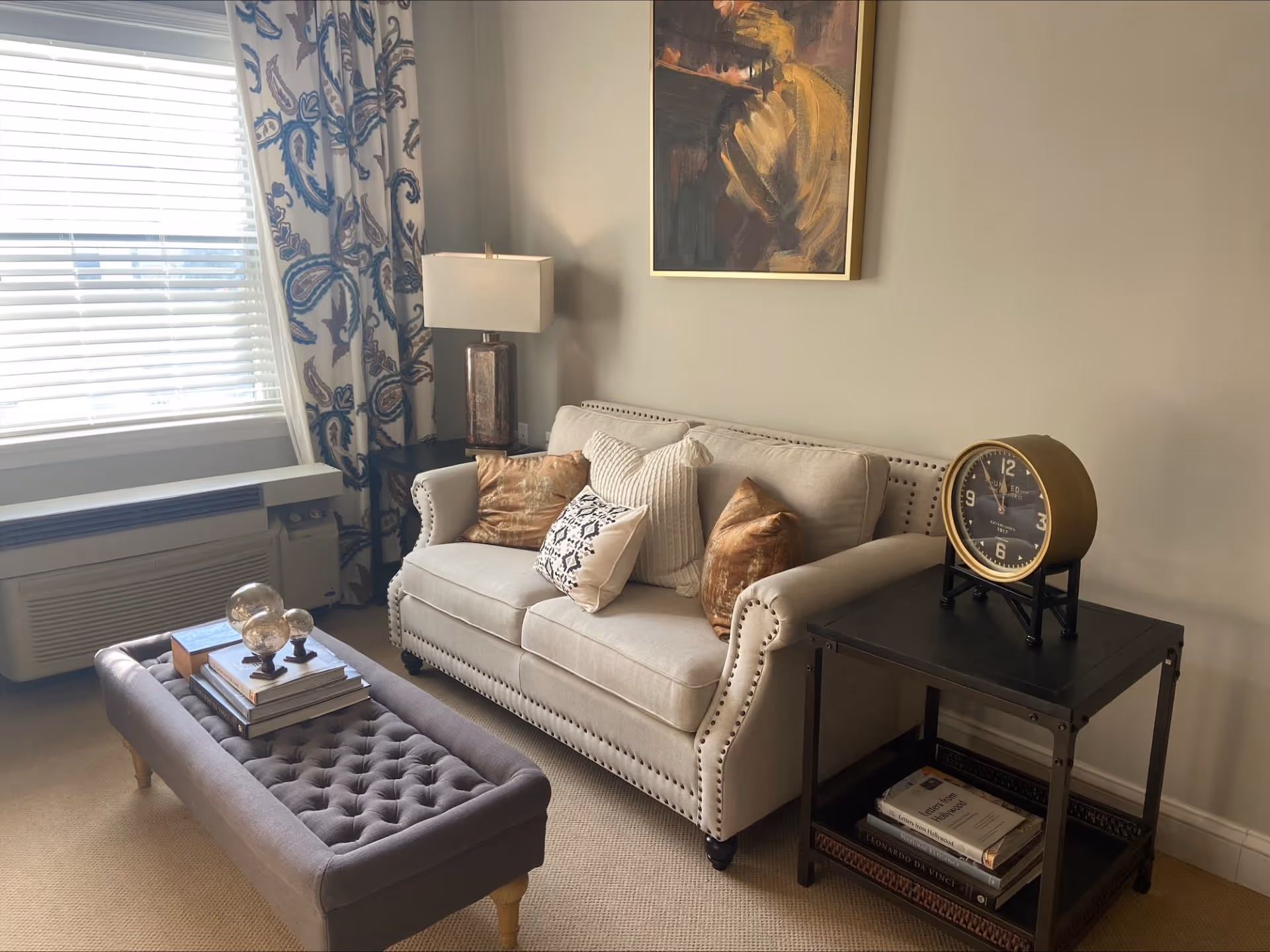 A cozy living room area with a beige upholstered sofa adorned with multiple decorative pillows, a dark wooden side table with a vintage-style clock on top, and a tufted gray ottoman with books and decorative glass spheres. A window with white blinds and patterned curtains allows natural light to fill the room. A framed abstract painting hangs on the wall above the sofa.