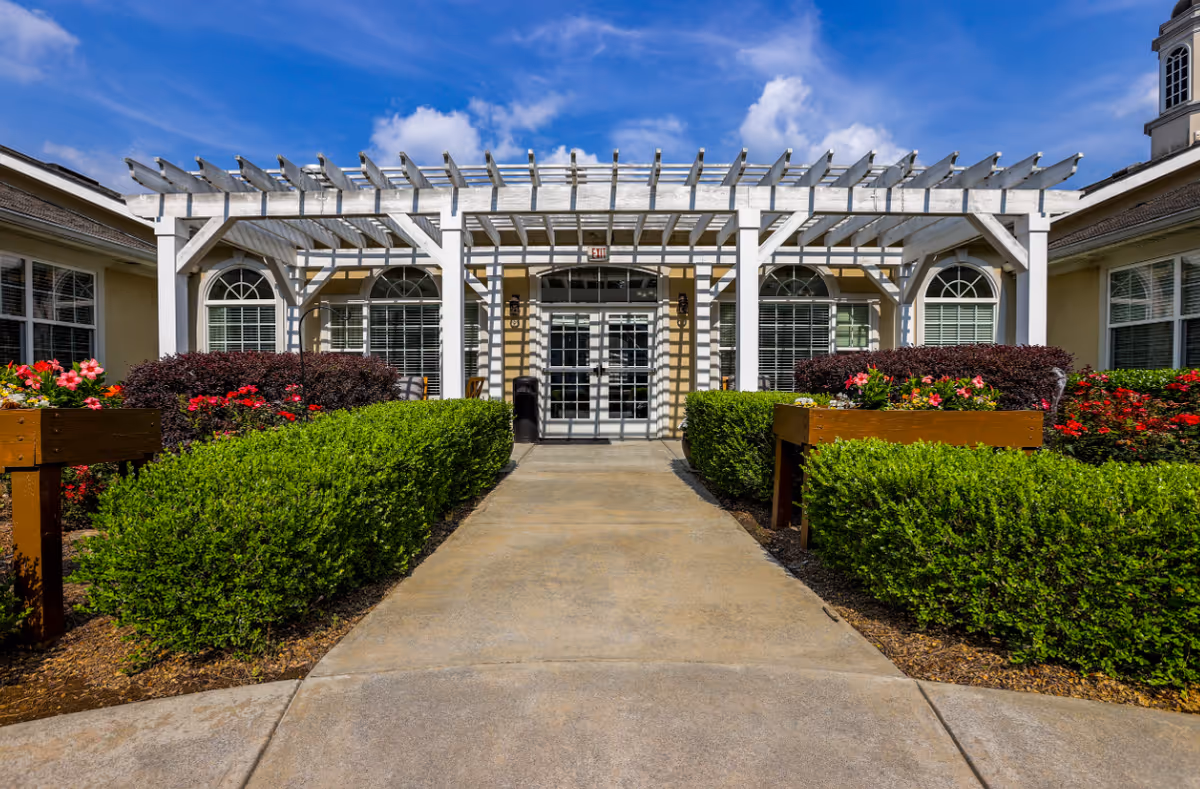 Front entrance of a senior living facility with a white pergola, arches, and manicured hedges.