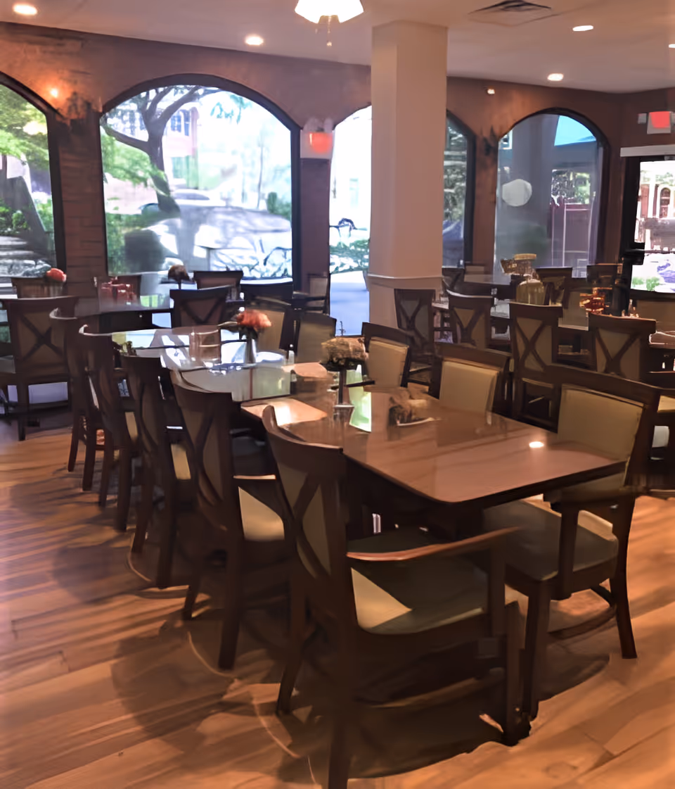 Spacious dining room with wooden tables and chairs, floral centerpieces, and large arched windows overlooking greenery.