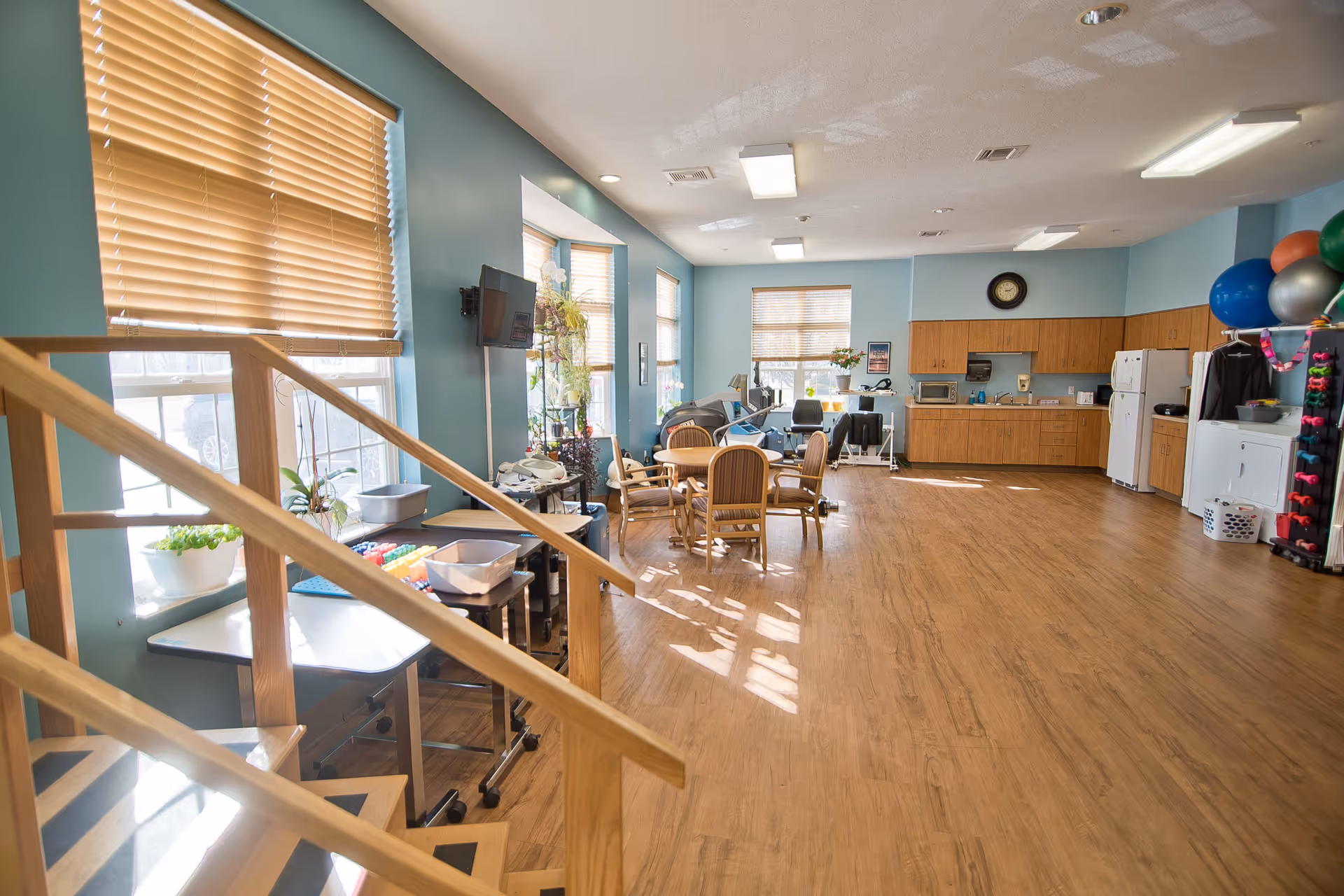A spacious, well-lit room with wooden flooring and light blue walls. The room features a small round table with four chairs in the center, a kitchenette area with wooden cabinets, a microwave, and a refrigerator on the right side. There are exercise balls and weights stored on racks near the kitchenette. Large windows with wooden blinds allow natural light to fill the room. A staircase with wooden railings is visible on the left side, and a wall-mounted TV is positioned near the windows.