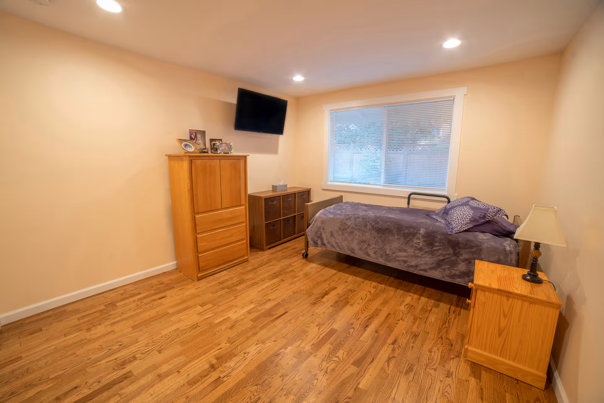 A sparsely furnished bedroom in a senior facility with bed, dresser, nightstand, and TV.