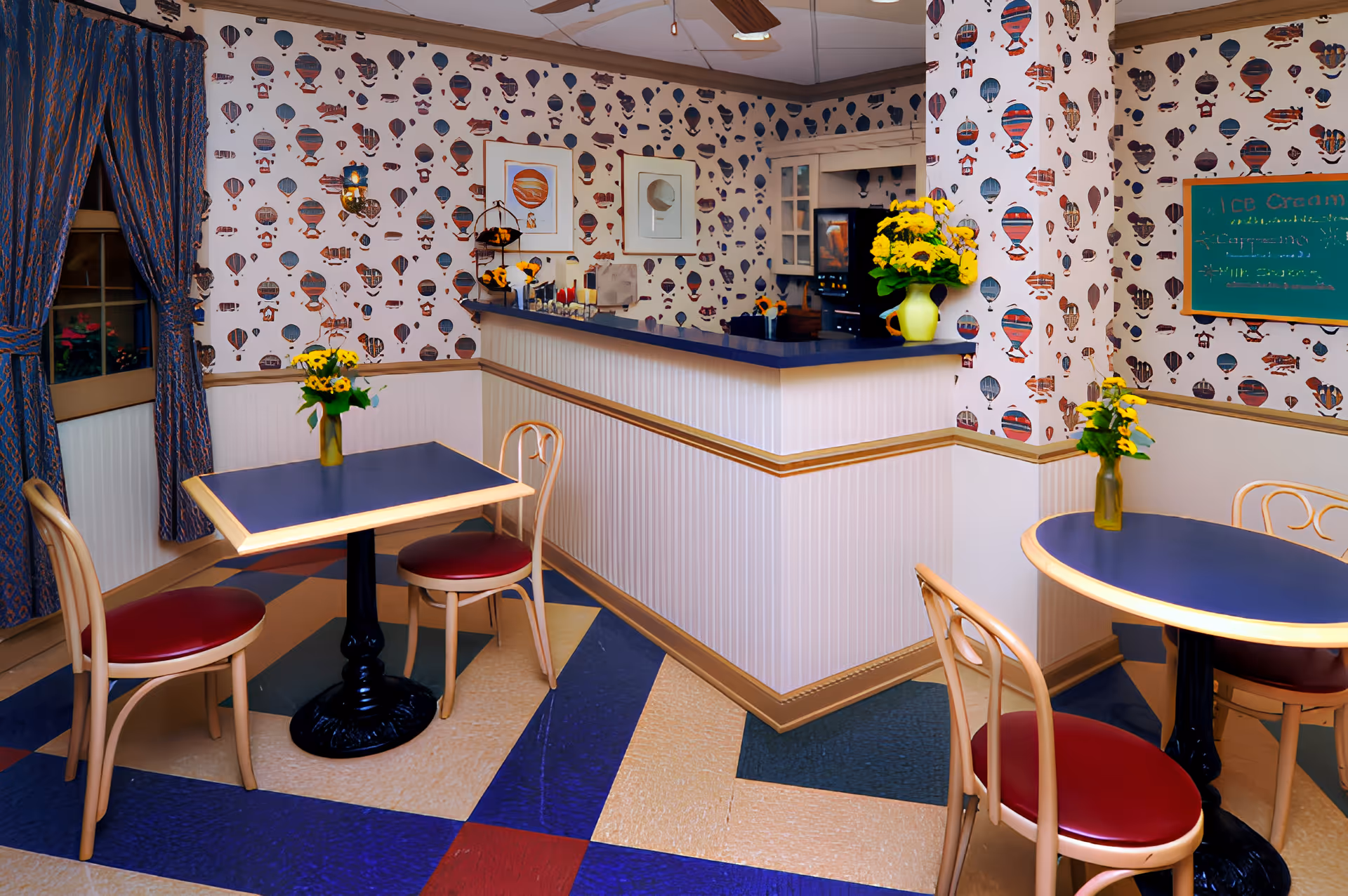 A cozy dining area with two small tables, each surrounded by two wooden chairs with red cushions. The tables have blue tabletops and small vases with yellow flowers. The walls are decorated with wallpaper featuring colorful hot air balloons and two framed pictures. A counter with a blue countertop and a vase of yellow flowers is visible, along with a window covered by blue patterned curtains. The floor has a geometric pattern in blue, beige, and red.