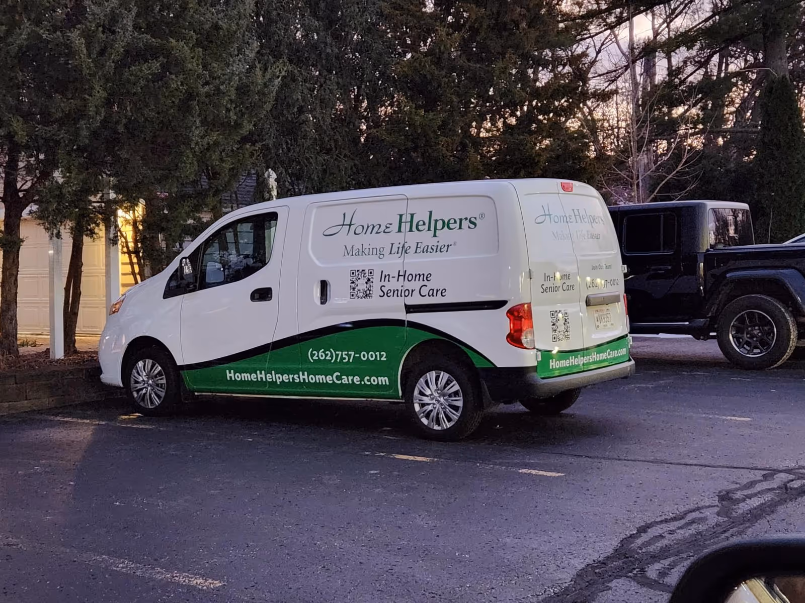 White Home Helpers van parked in a lot with trees and a dark pickup nearby.