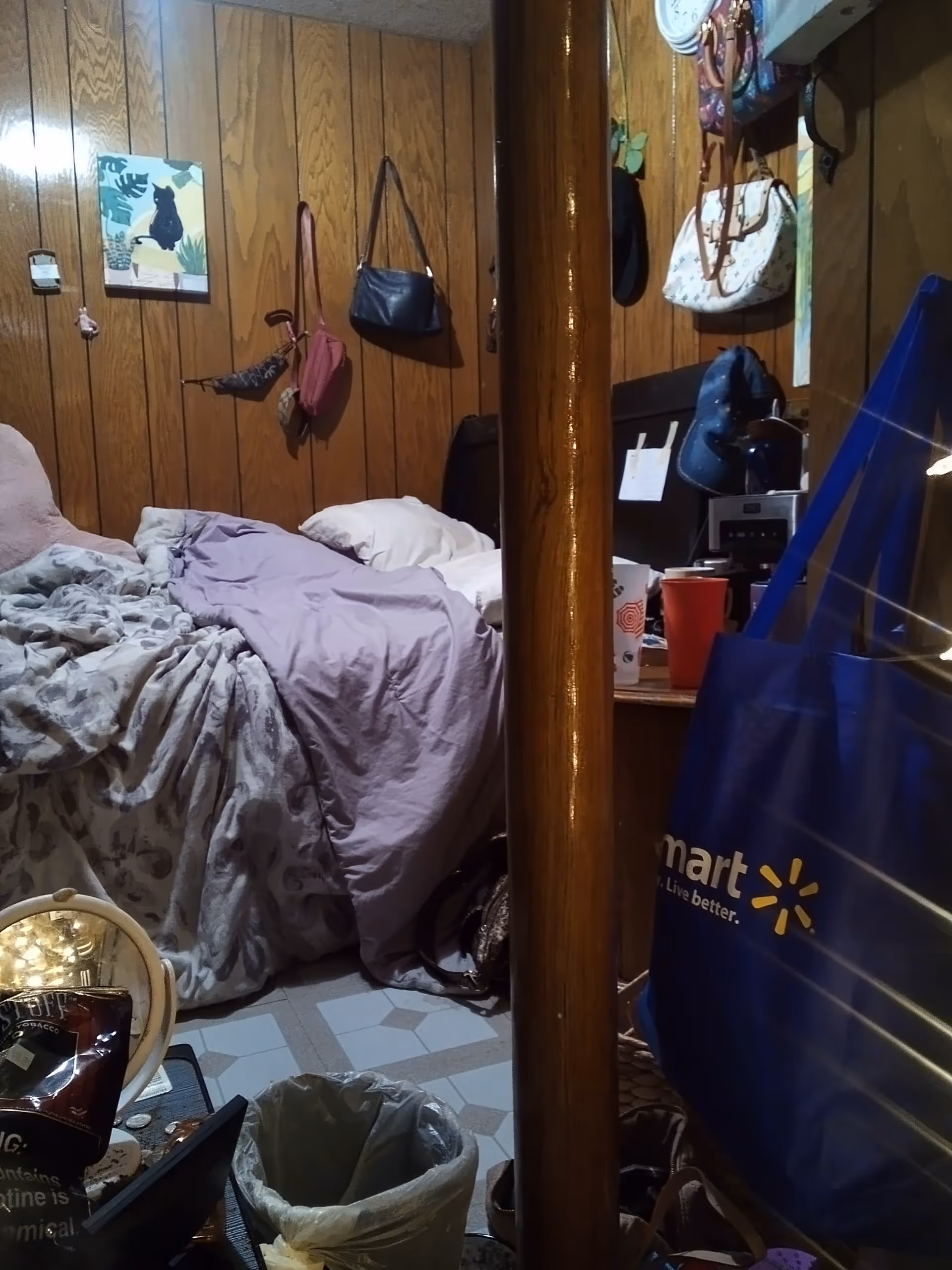 Cluttered senior living bedroom with a bed, wood-paneled walls, and hanging bags.