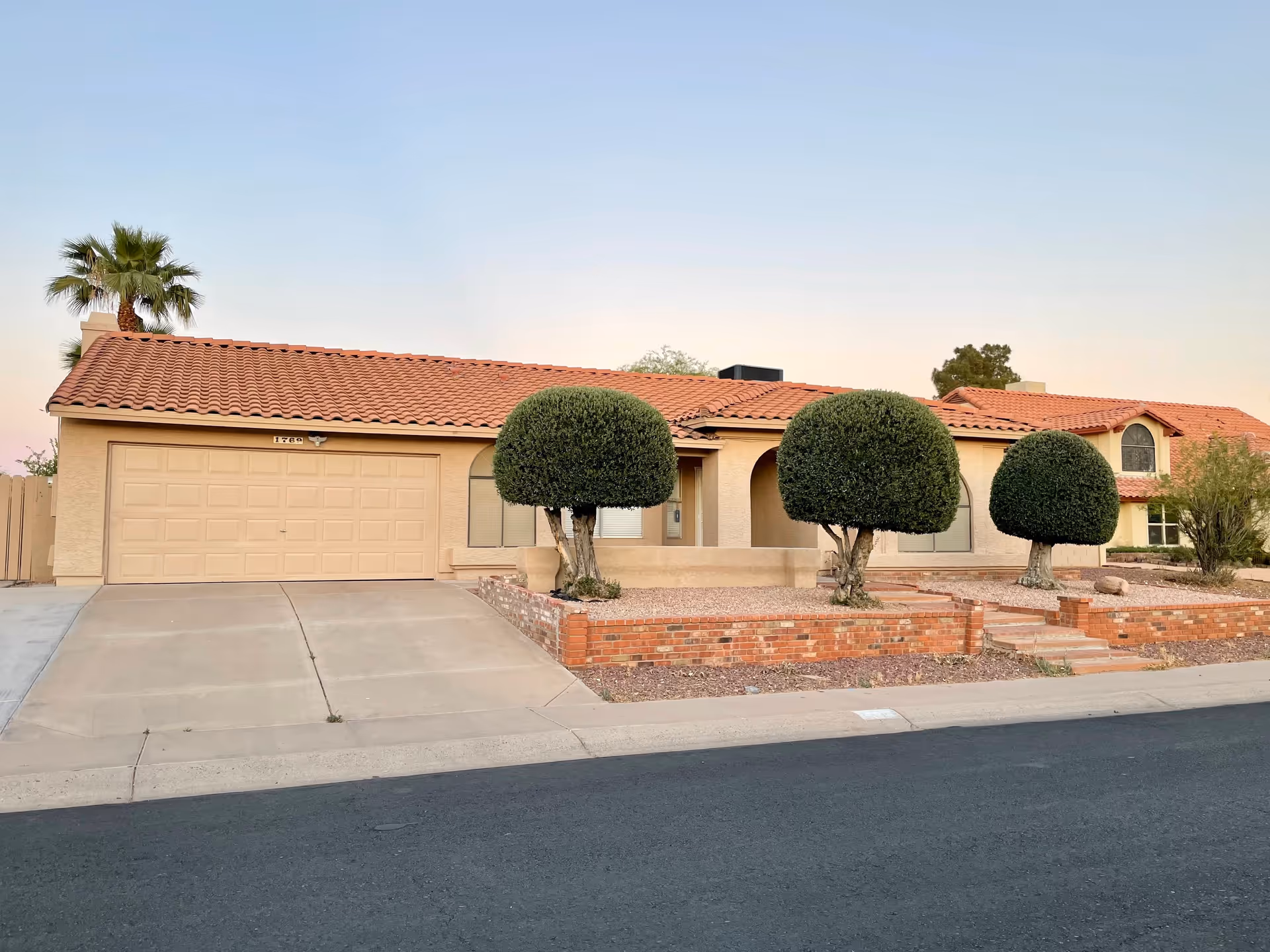 Single-story stucco house with a red tile roof, two-car garage and a front yard featuring three rounded topiary trees and brick planters.