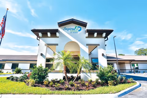 Exterior front entrance of the Inspired Living Tampa senior living building with palm trees, landscaped beds, and an American flag.