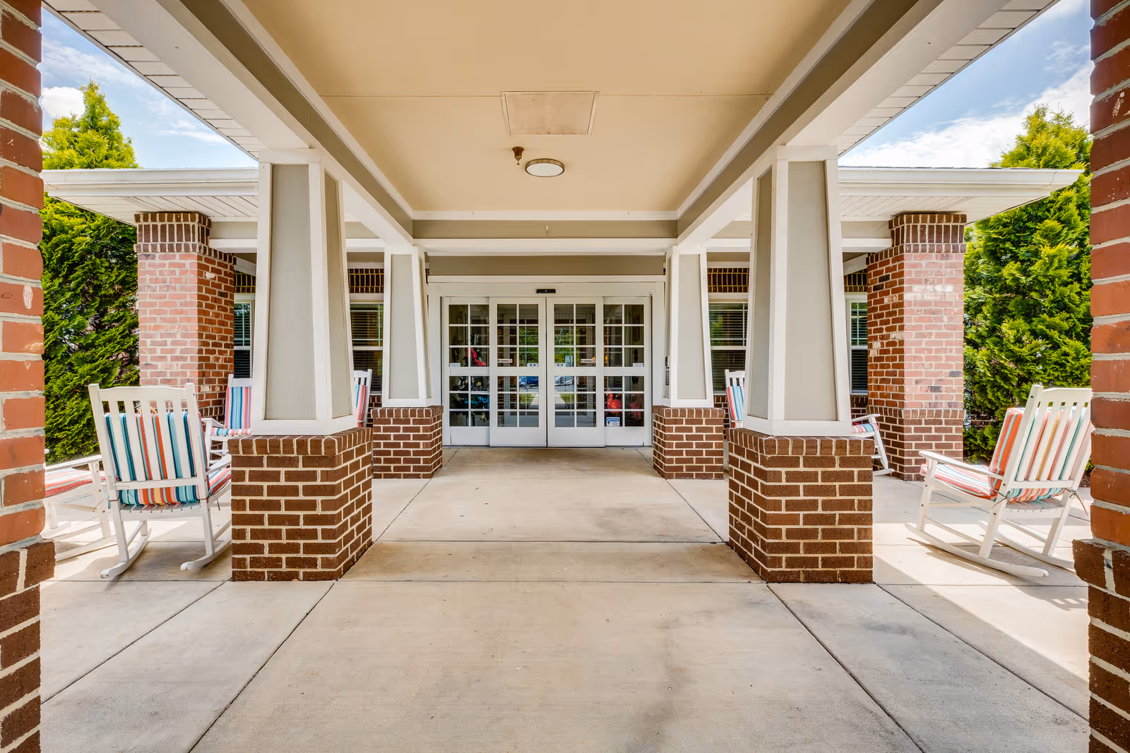 Covered entrance area of a building with brick columns and white rocking chairs with striped cushions on either side. Double glass doors are visible at the far end, with greenery and blue sky in the background.
