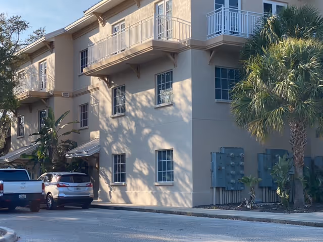 Exterior view of a beige senior living building with parked cars and palm trees.