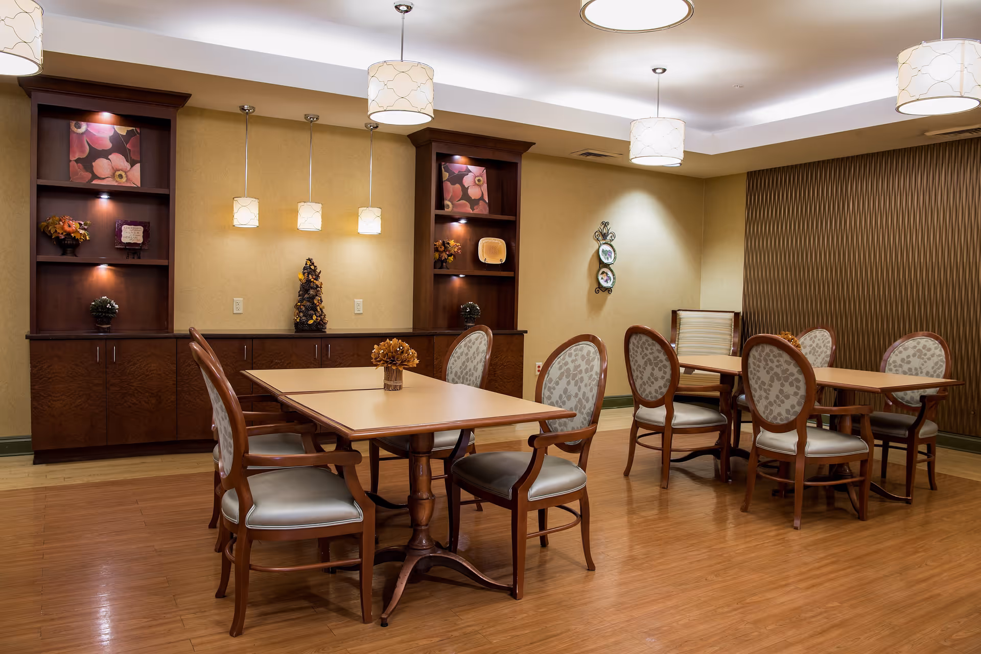 A well-lit dining room with wooden floors, two rectangular tables, and several upholstered chairs arranged around them. The walls are decorated with built-in wooden shelves holding floral decorations and artwork. Pendant lights hang from the ceiling, providing warm lighting.