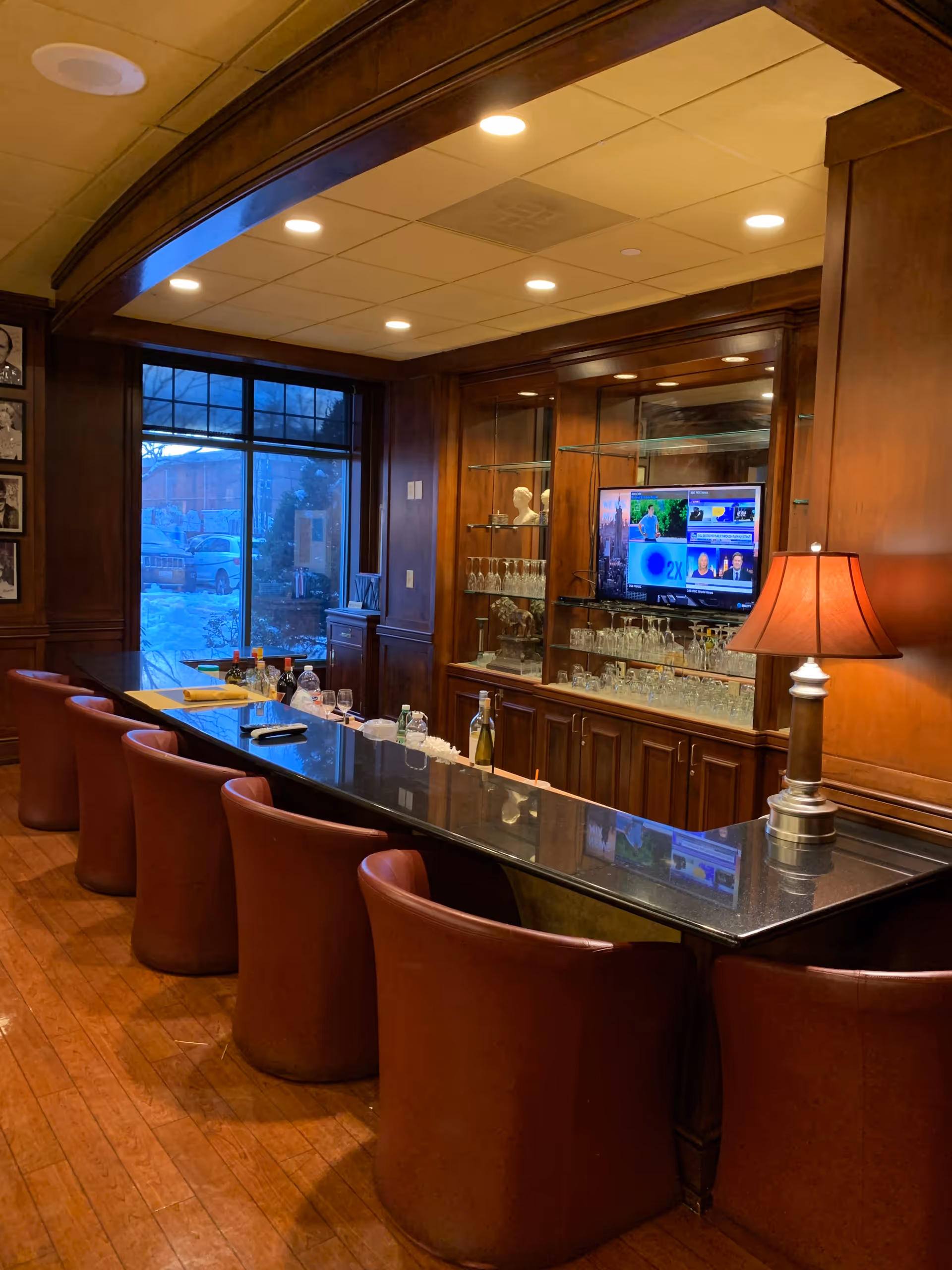 Interior view of a bar area with a long black countertop and six brown leather chairs. Behind the bar is a wooden cabinet with glass shelves holding glassware and decorative items. A flat-screen TV is mounted on the cabinet displaying a news channel. A table lamp with a red shade is placed on the bar counter. Large windows show a snowy outdoor scene.