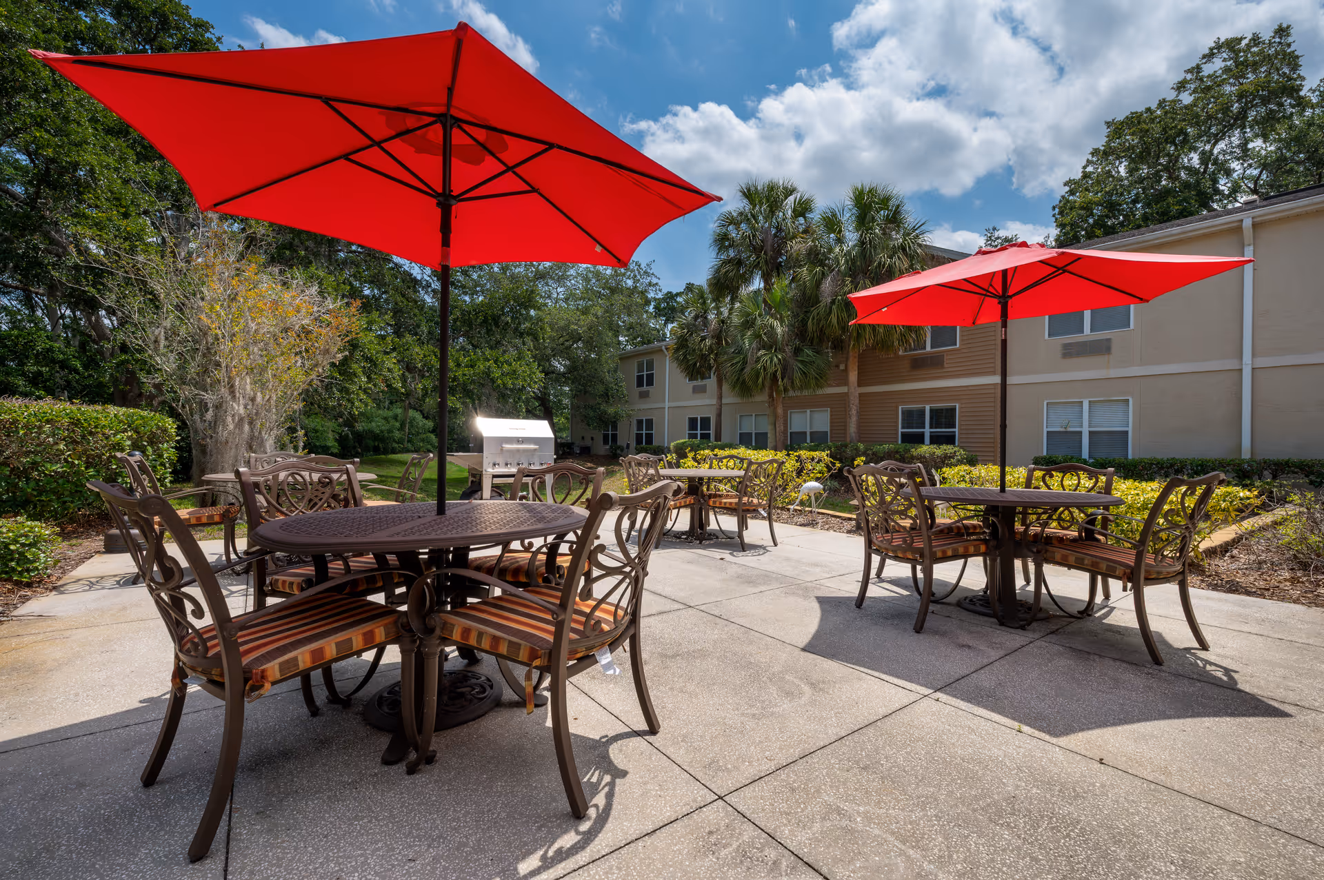 Outdoor patio area with two round metal tables, each surrounded by four chairs with striped cushions. Each table has a large red umbrella providing shade. In the background, there are palm trees, bushes, and a two-story building under a partly cloudy sky.
