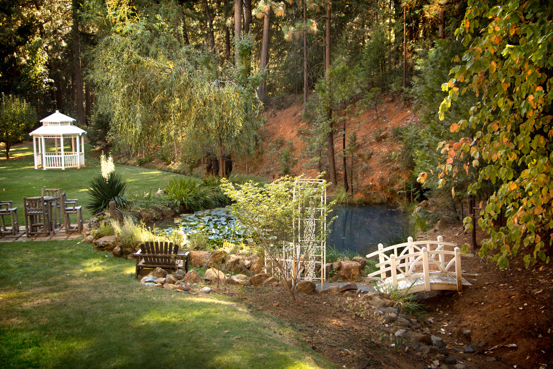 A landscaped outdoor garden with a pond, white bridge, gazebo, and benches.