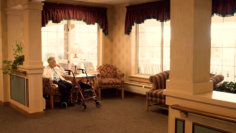 An elderly woman sitting on a patterned armchair next to a walker in a warmly lit common area with large windows, floral wallpaper, and additional seating including a striped sofa and another armchair. A table lamp is placed near the window, and there are plants on the left side.