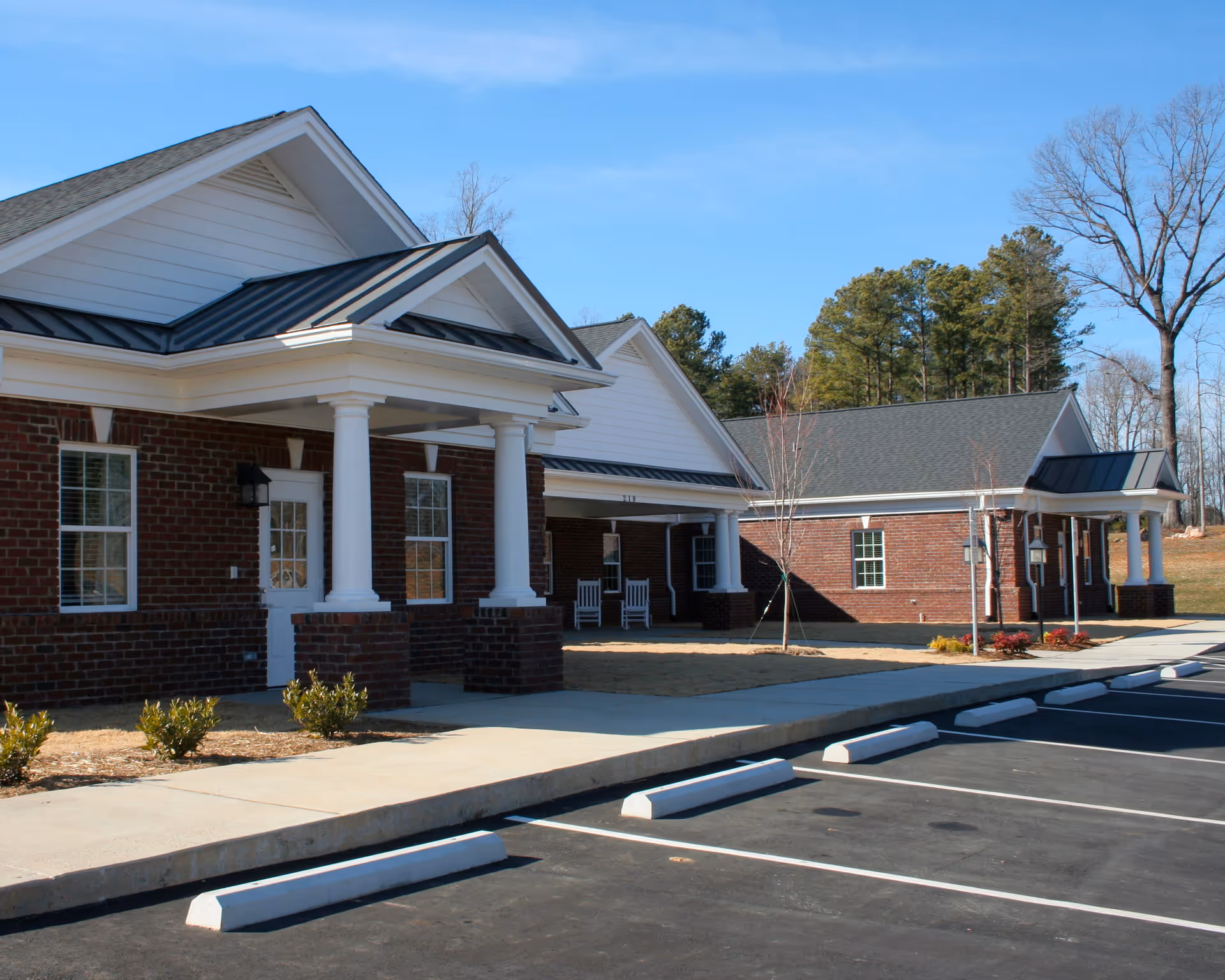 Exterior view of Village Lake Inn of Siler City showing a brick building with white columns and a covered entrance. There are two white rocking chairs on the porch, small shrubs planted along the sidewalk, and a parking lot with marked spaces in the foreground. Trees and a clear blue sky are visible in the background.