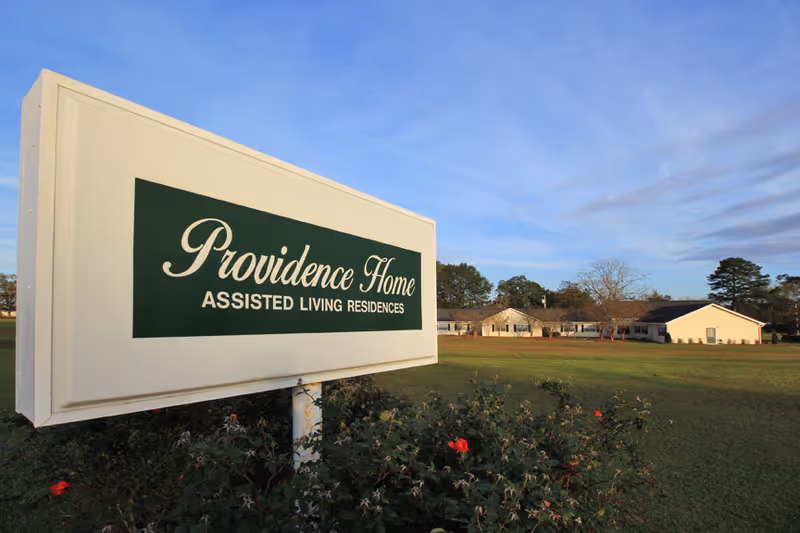A sign reading 'Providence Home Assisted Living Residences' on a lawn with the facility buildings visible in the background.