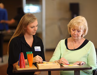 A young female caregiver or staff member sitting at a table with an elderly woman, engaging in a board game or activity involving colored pieces in a well-lit indoor setting.