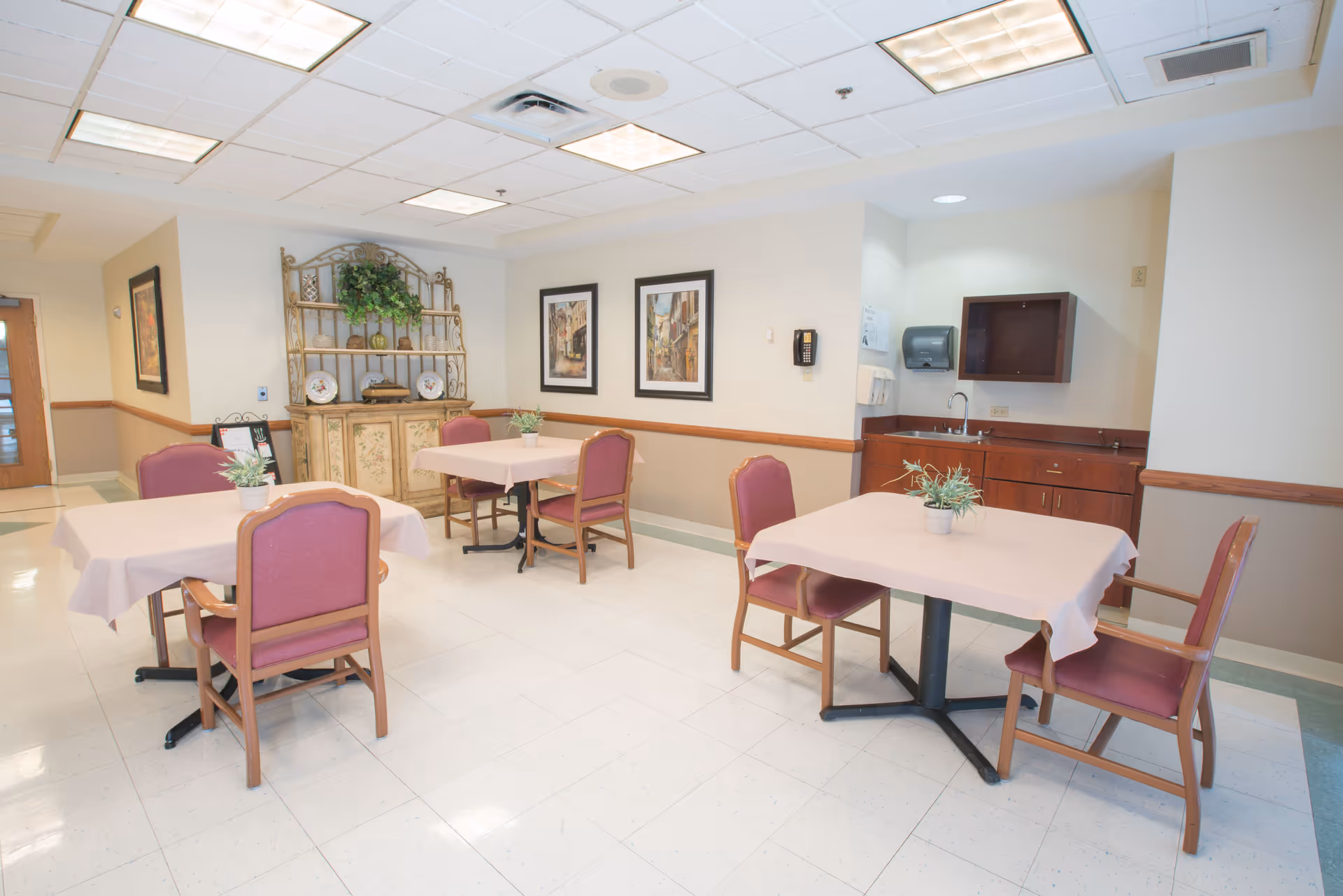 A small dining area with three tables covered in light pink tablecloths, each surrounded by wooden chairs with pink cushions. There are small potted plants on each table. The room has a white tiled floor, beige walls with a wooden chair rail, and a ceiling with fluorescent lights. On one side, there is a wooden cabinet with a sink, paper towel dispenser, and a wall-mounted phone. The opposite wall features a decorative wooden shelf with plants and plates, and two framed pictures.