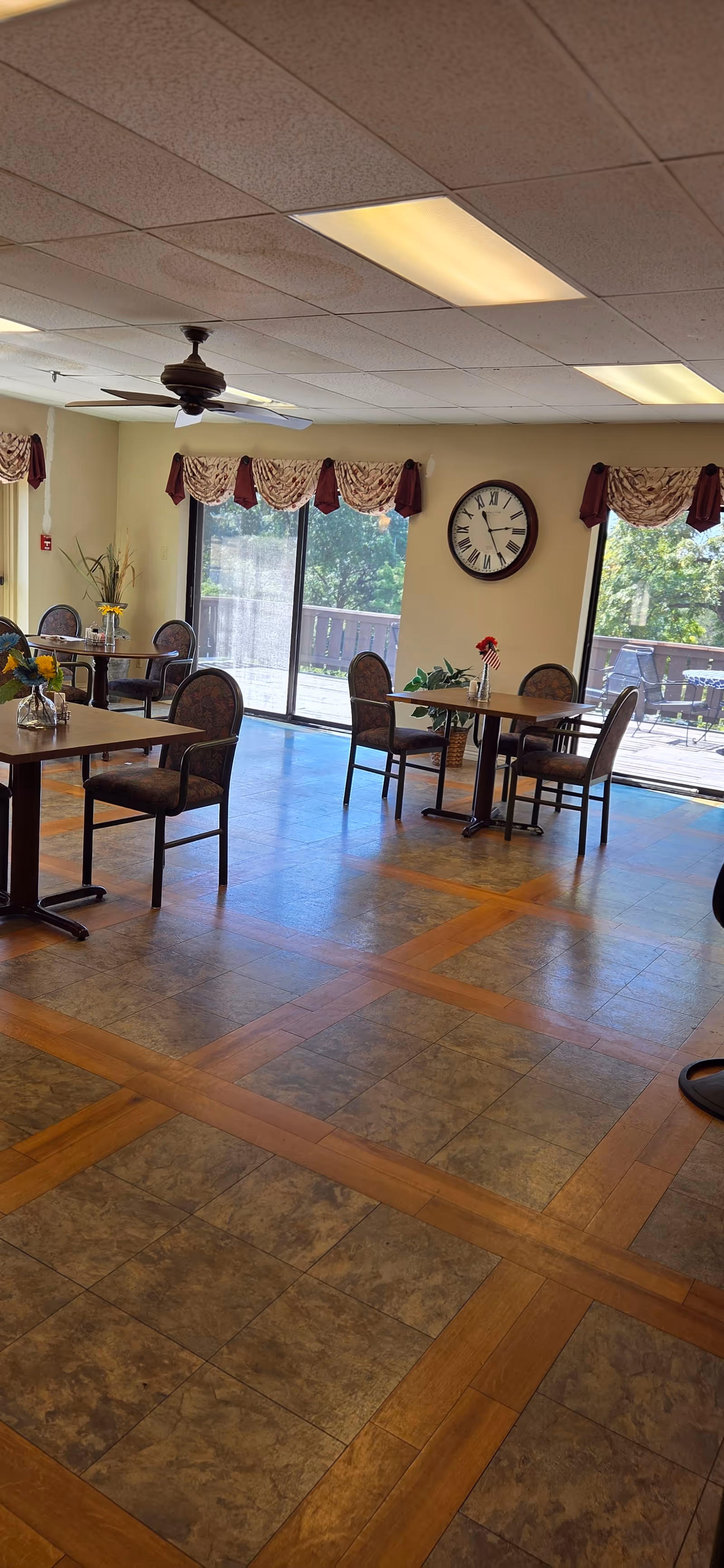 Marymount Manor dining room interior with round tables, chairs, clock, windows, burgundy curtains.