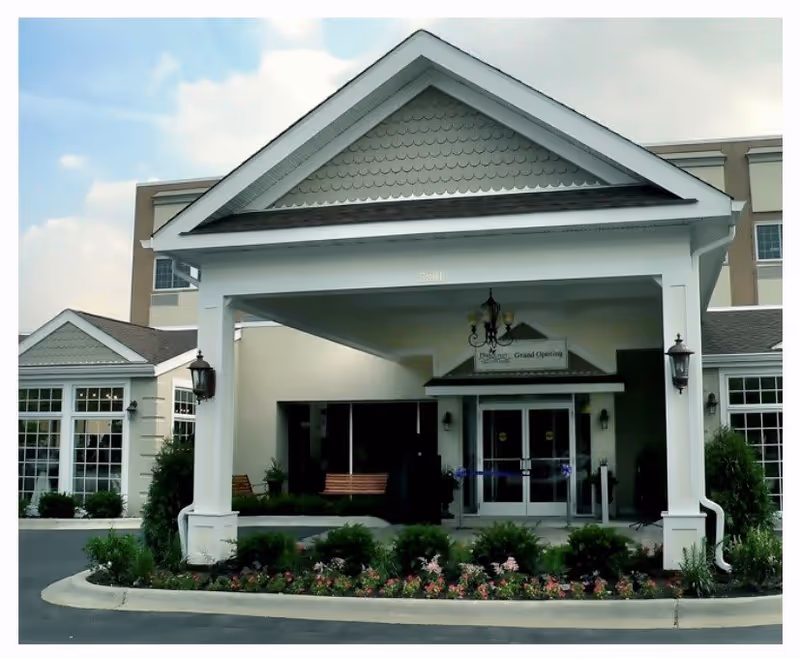 Front entrance of Plum Creek Supportive Living facility featuring a covered drop-off area with white pillars, a chandelier hanging from the ceiling, and a flower bed with various plants and flowers in front. The building has large windows and beige exterior walls.
