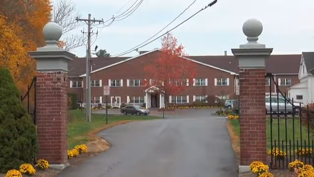 Brick senior living facility entrance with stone pillars, gate, and fall-colored trees.