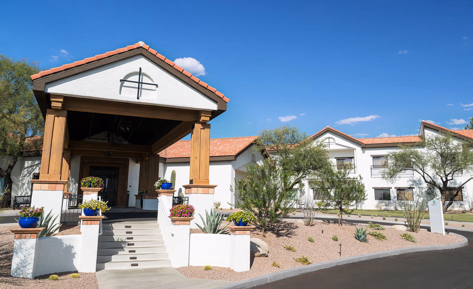 Front entrance of a white stucco senior living building with a covered porte-cochere, potted flowers, and desert landscaping under a clear blue sky.