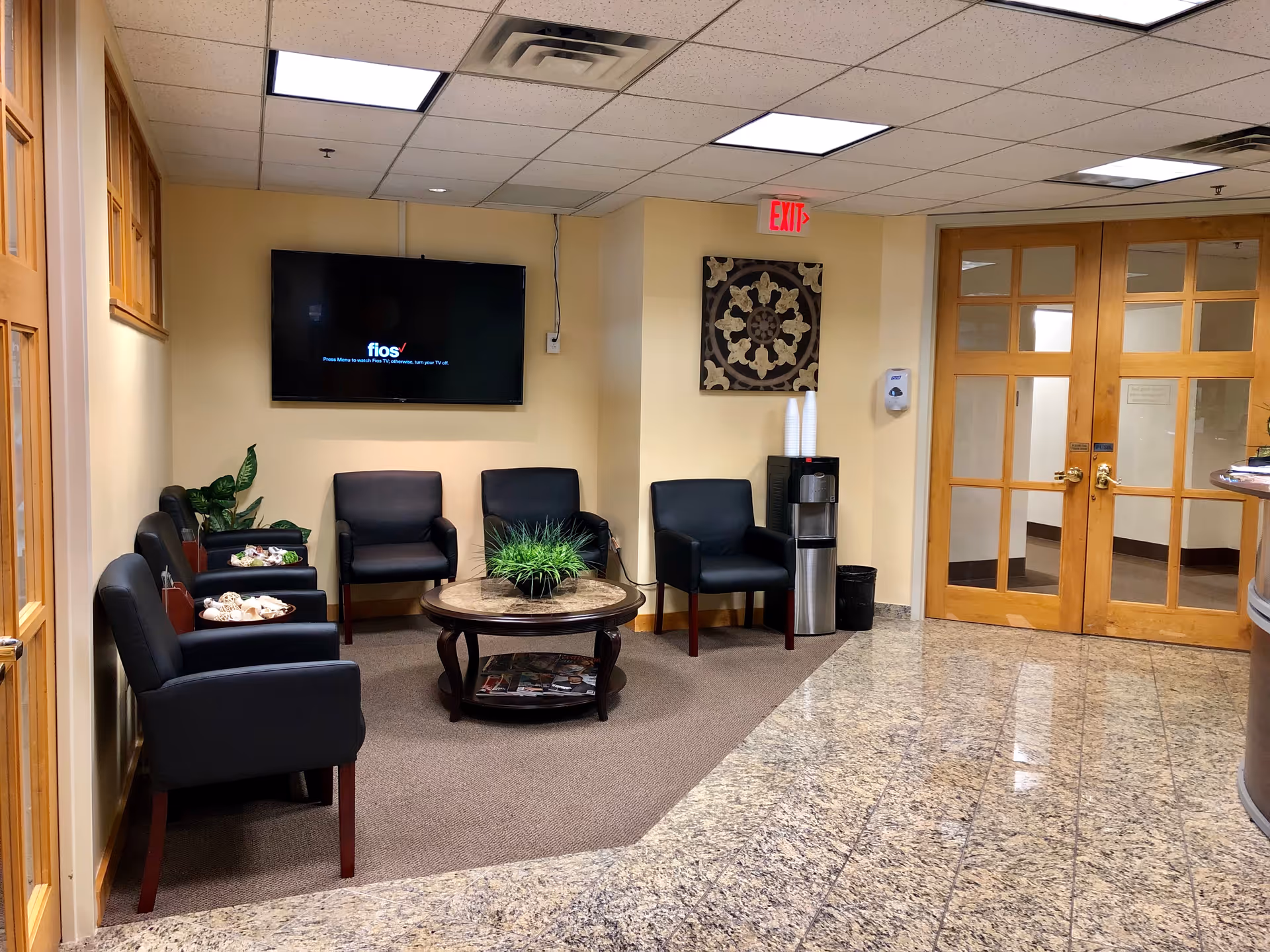 Lobby waiting area with black chairs, a coffee table, TV, plants, and glass doors.