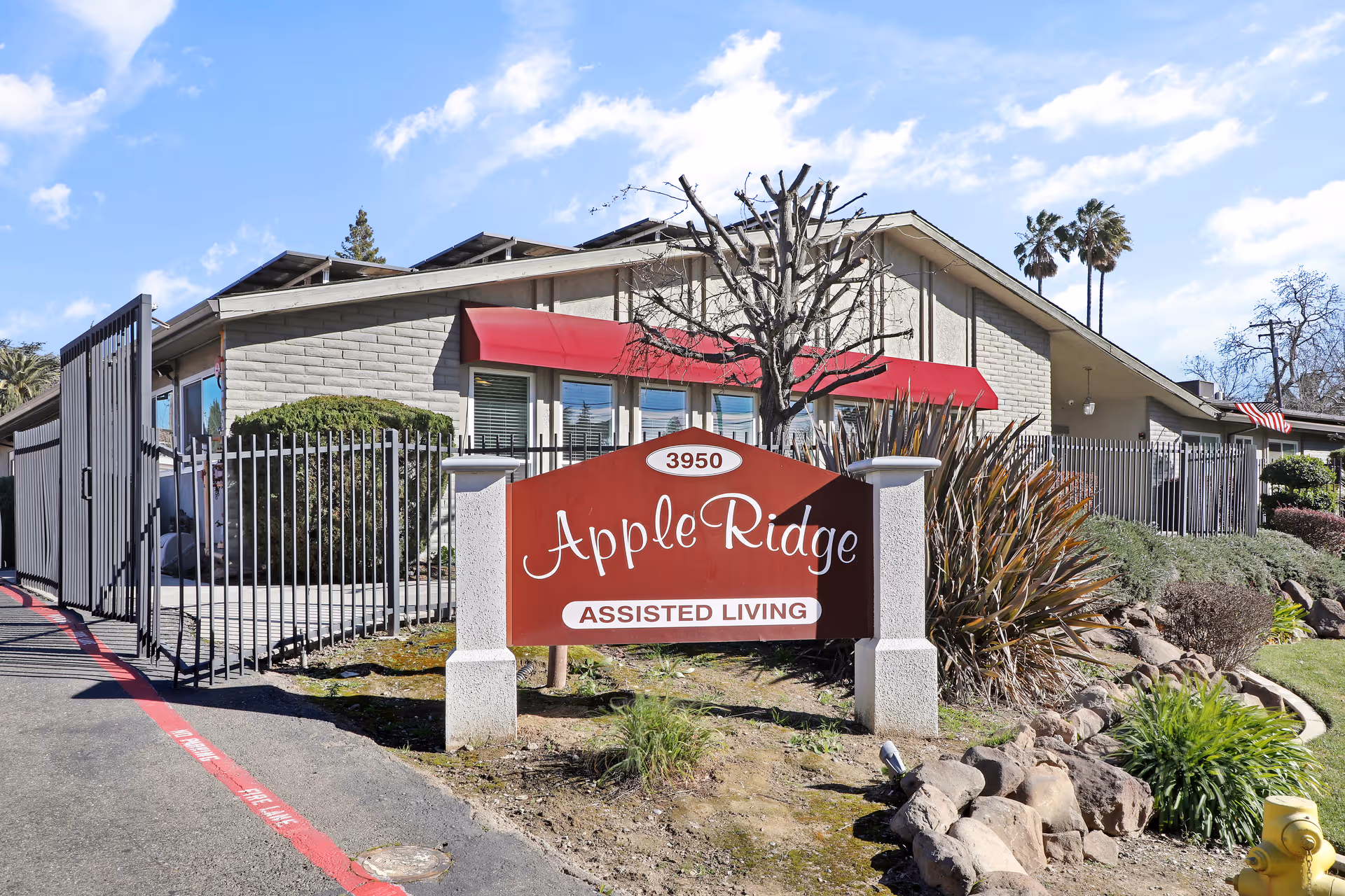 Exterior view of Apple Ridge Assisted Living facility showing a red sign with white text, a gated entrance, a building with red awnings, and landscaping with bushes and a tree under a partly cloudy blue sky.