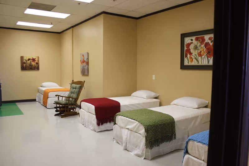Shared dorm-style bedroom in a senior care facility with multiple single beds and colorful throws.