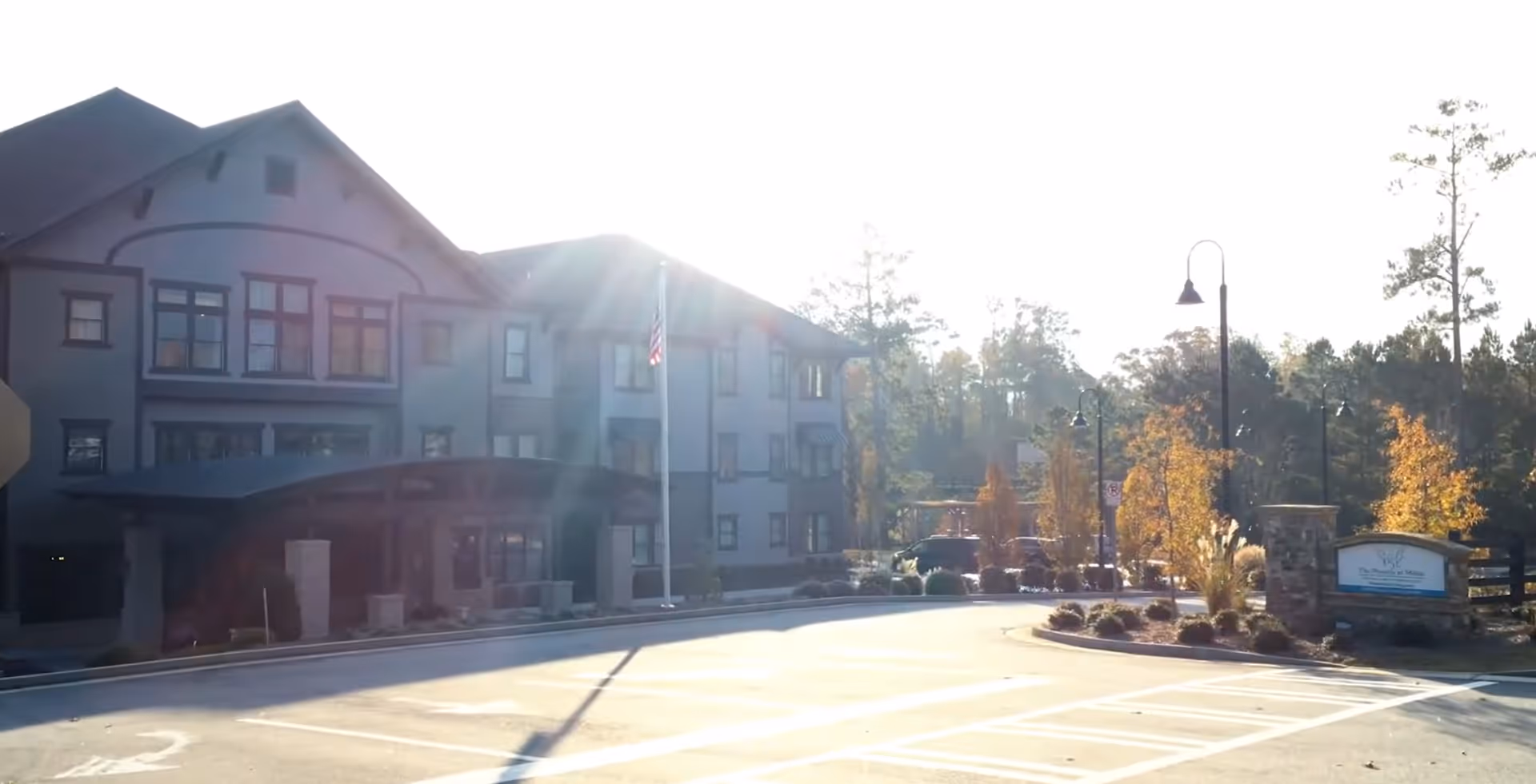 Exterior front entrance of a multi-story senior living building with a drive-up porte-cochère, flagpole, and landscaped roundabout.