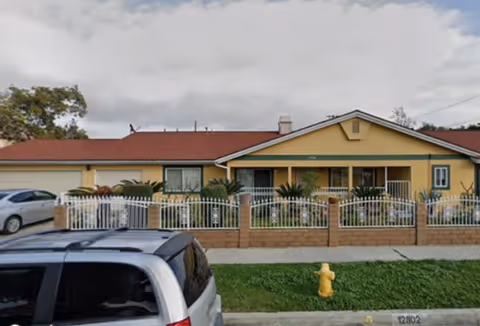 Front exterior of a single-story yellow facility with a brick fence and parked cars.