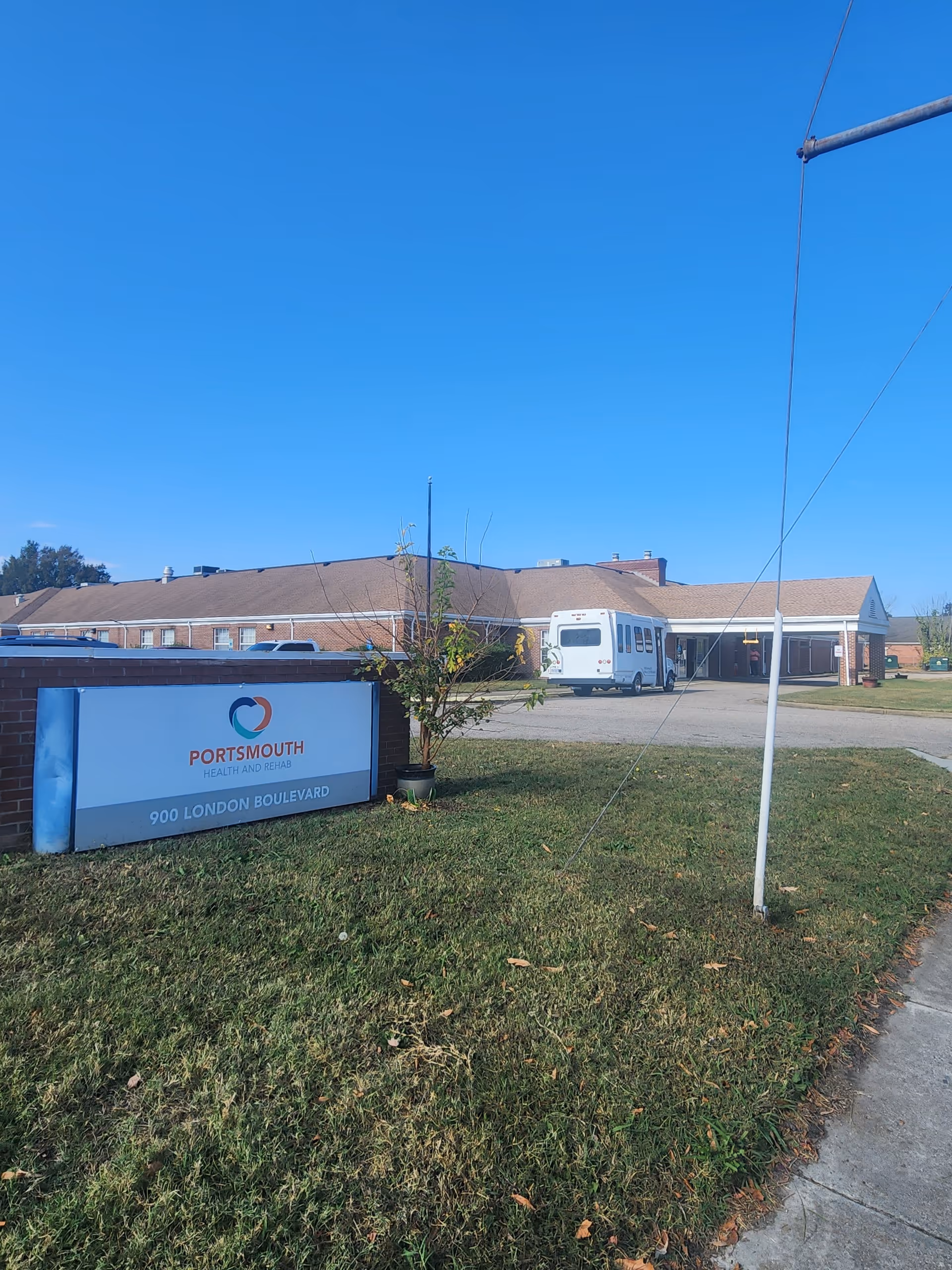 Front exterior of Portsmouth Health and Rehab with a sign on the lawn and a shuttle van parked by the building under a clear blue sky.