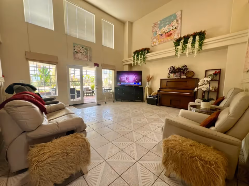 A bright living room with high ceilings and large windows. The room features two beige recliner sofas, two furry ottomans, a piano with decorative plants on top, a flat-screen TV on a black stand, and a glass door leading to an outdoor patio. The walls are decorated with floral paintings and hanging greenery.