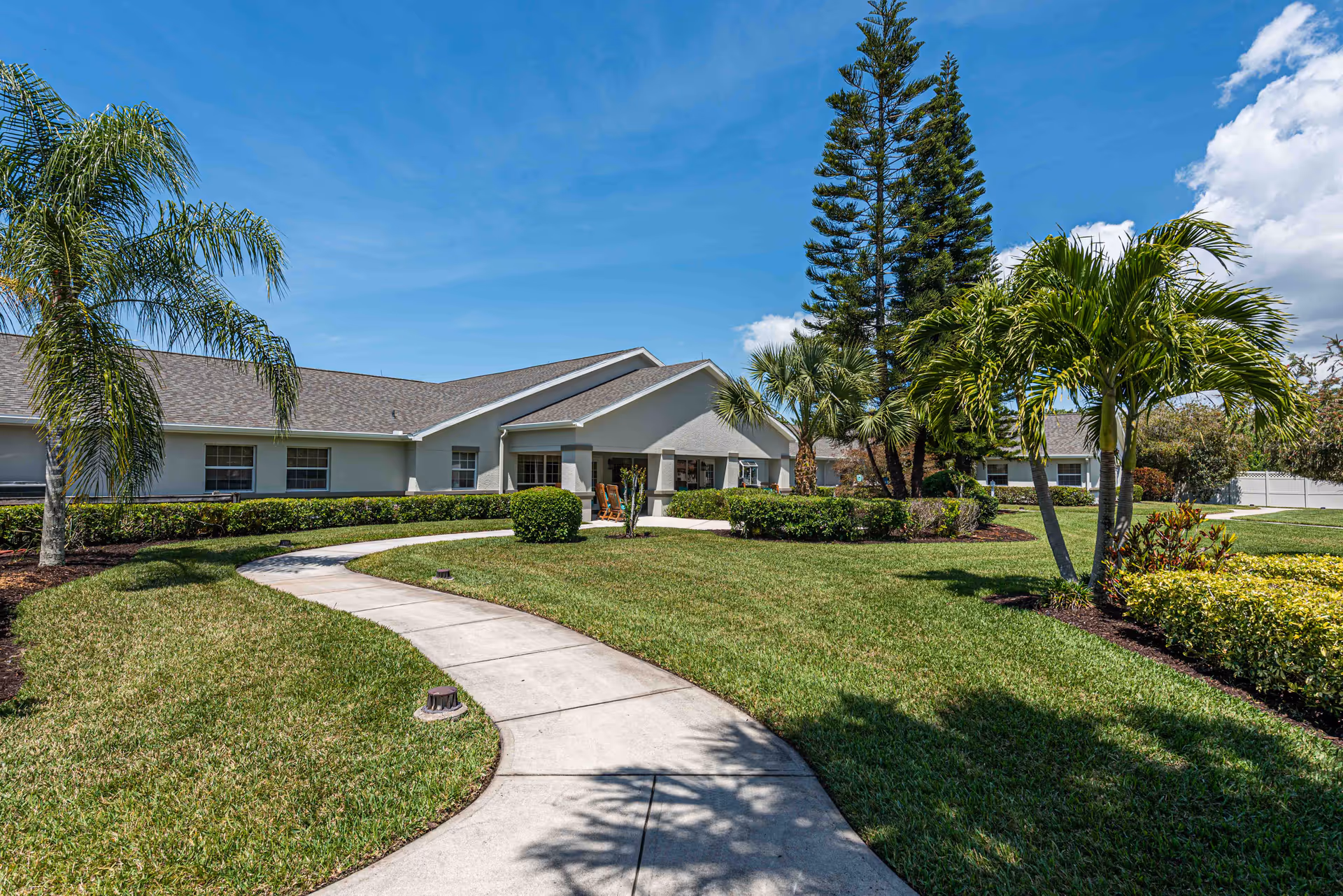 Sunny exterior of Arden Courts Sarasota with a curved walkway, manicured lawn, and palm trees.