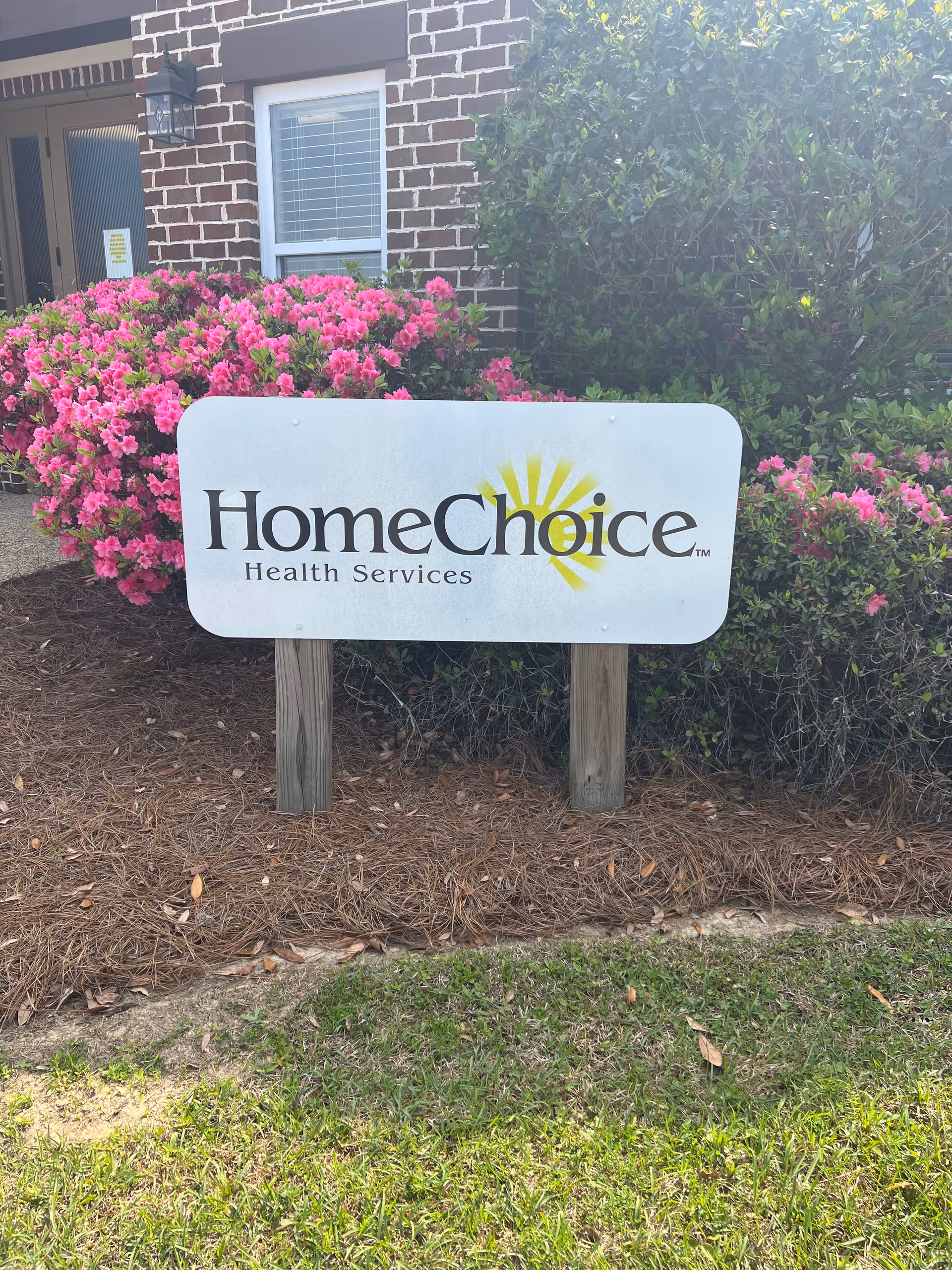Outdoor view of a brick facility entrance with a large HomeChoice sign and pink flowering shrubs.