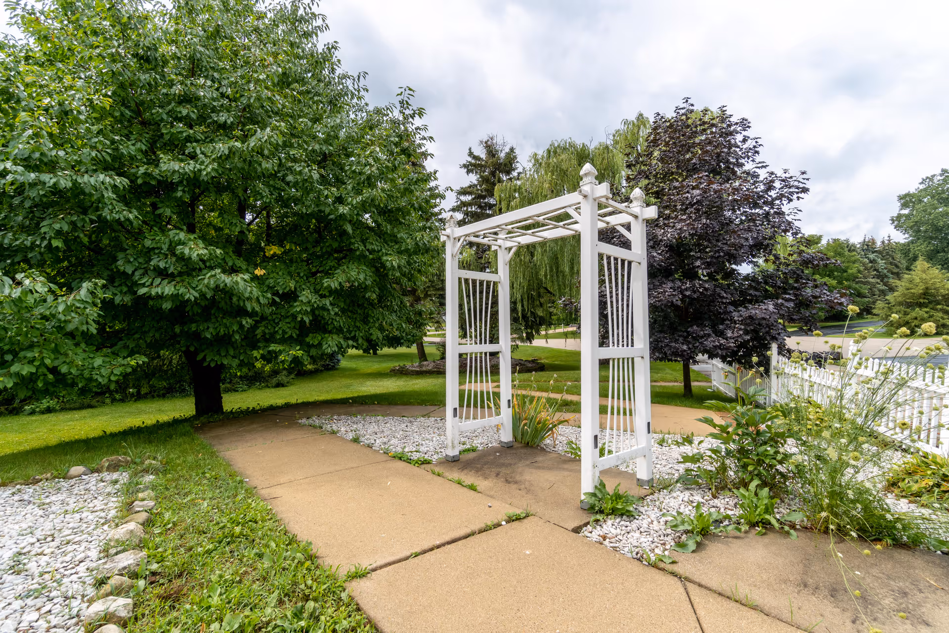 White garden trellis over a paved path surrounded by green trees and shrubs.