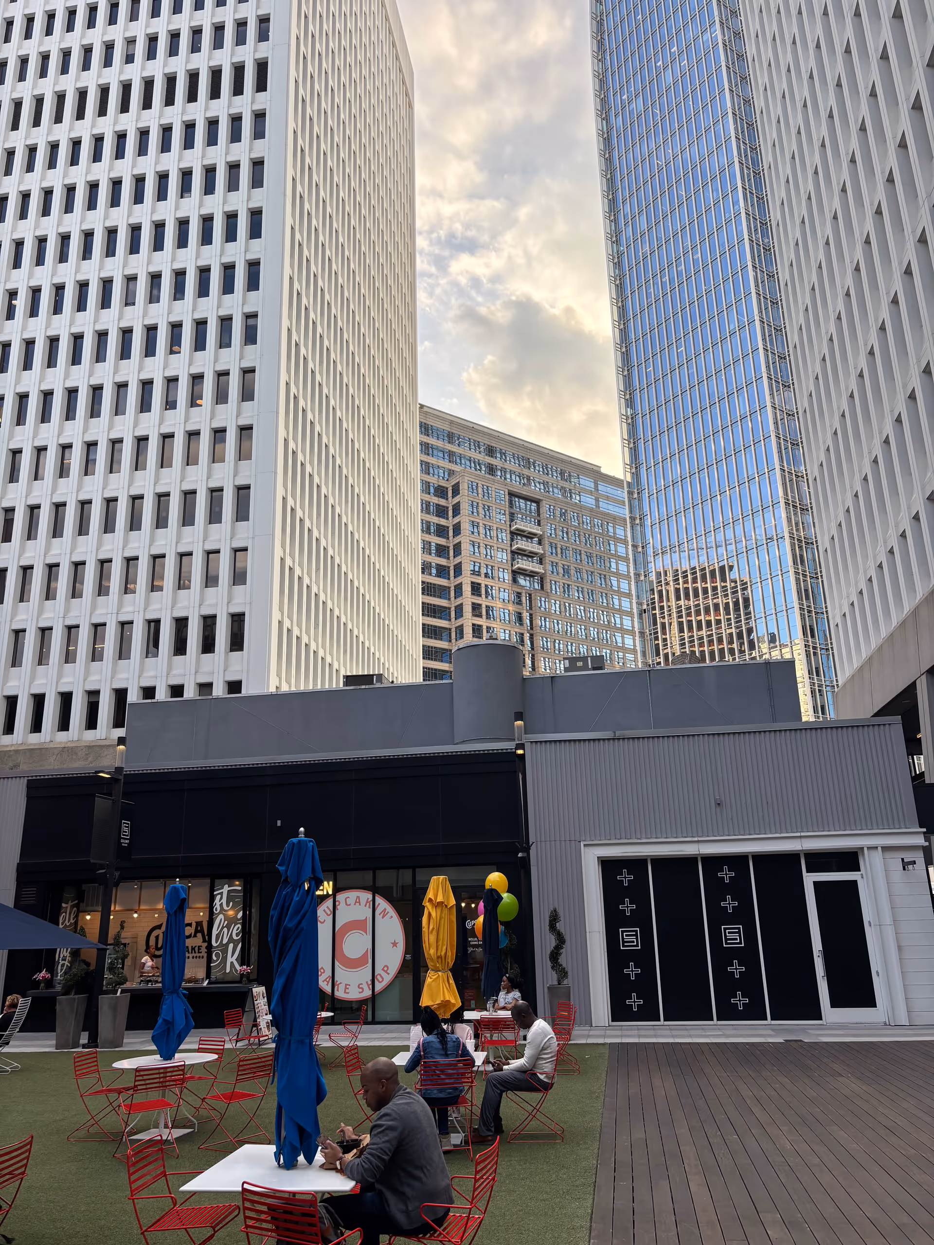 Outdoor courtyard with red chairs, umbrellas, and a few people among tall surrounding buildings.