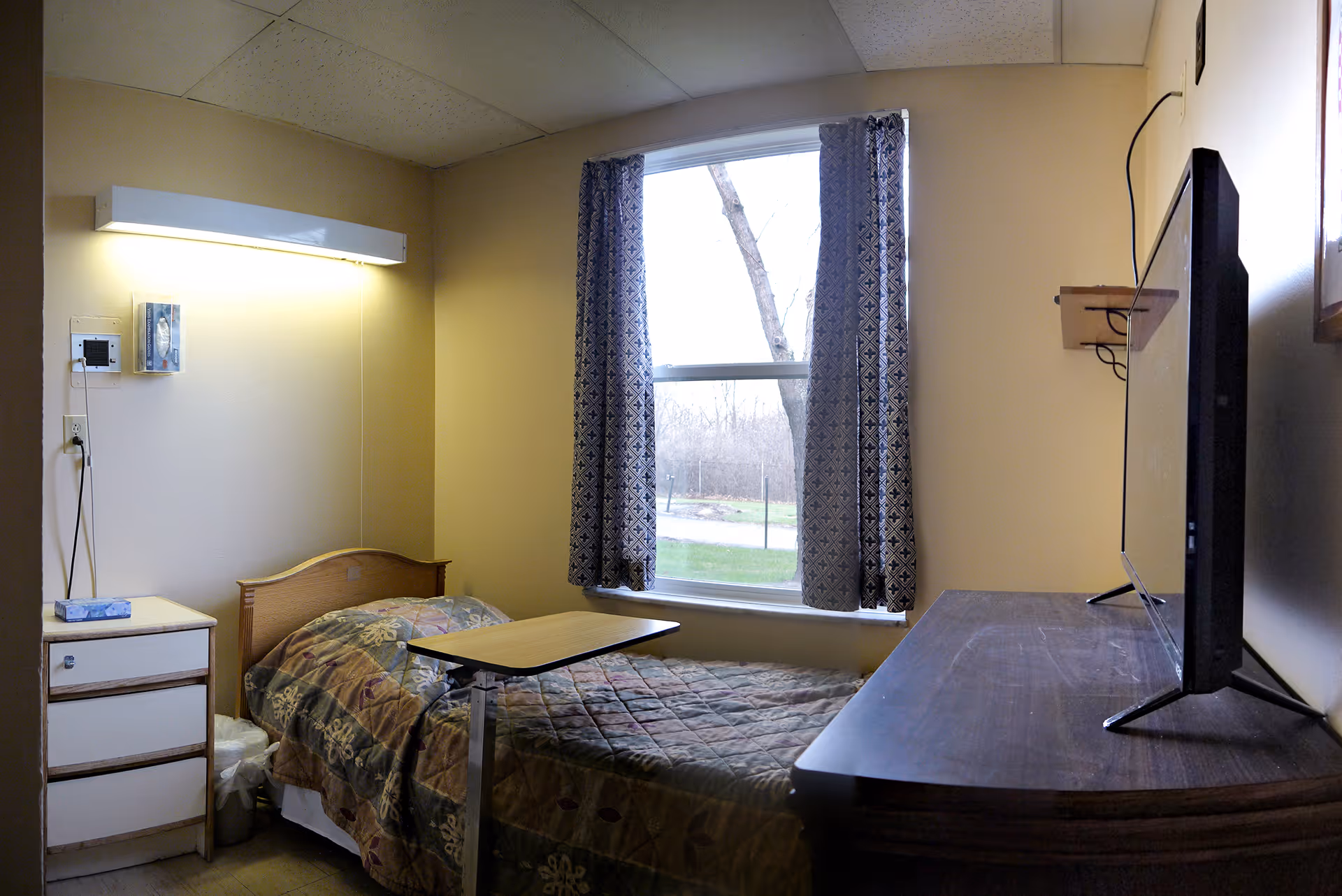 A small bedroom in a senior living facility with a single bed covered with a patterned quilt, a bedside table with a box of tissues, a window with patterned curtains, and a flat-screen TV on a wooden dresser.