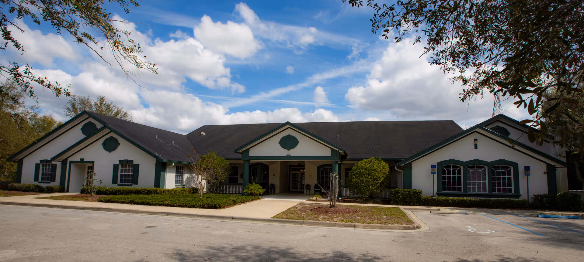 Single-story senior living facility exterior with a central entrance, green shutters, and surrounding landscaping.